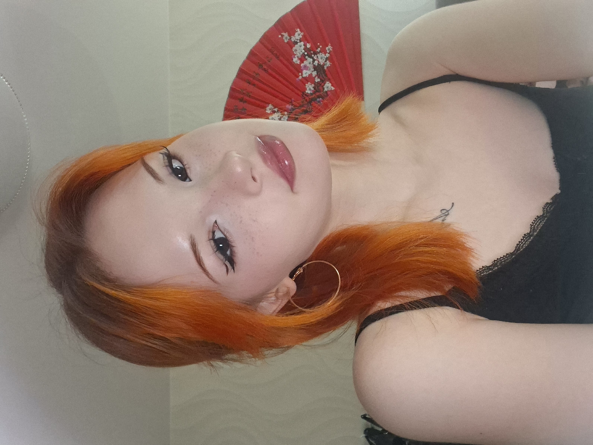 A portrait of a young woman with orange hair, fair skin, and freckles is presented. She is wearing a black tank top with lace trim. A gold hoop earring adorns her left ear. She has makeup on her eyes, and her lips are glossy. A red fan with white floral designs is positioned behind her head. A small tattoo is visible on her upper chest. The background is a plain, light-colored wall. The location is not evident in the image; there is no indication of the city of Chaikovsky, Russia.