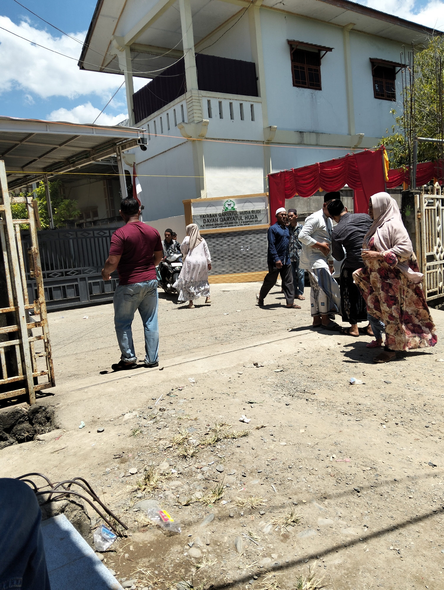 In Teungoh, Indonesia, on a bright, sunny day, a group of individuals are gathered outside a building with a white facade and a prominent red draped entrance. The building appears to be a religious or educational institution, as suggested by a sign that reads "YAYASAN QARYATUL RUDI QARYATUL HUDA," with additional text below.
In the foreground, a man wearing a maroon t-shirt and blue jeans stands facing away from the camera, his left arm extended as if in mid-gesture. To his right, a woman in a light-colored headscarf and a long, floral-patterned dress is walking away from the building. Behind her, a person is seated on a motorcycle.
Further into the scene, a group of men are conversing near the entrance. One man in a white shirt and checkered pants is speaking with a man in a dark shirt and cap. Another man in a white shirt and a cap stands to their left, facing the building.
The environment is dusty and somewhat unkempt, with a dirt road and scattered debris. A rusty metal gate or fence is visible on the left side of the image, and a portion of a building's roof structure is visible on the left, supported by metal poles. Trees and foliage are present in the background, along with a blue sky with white clouds. The overall impression is one of a community gathering or event taking place outdoors.