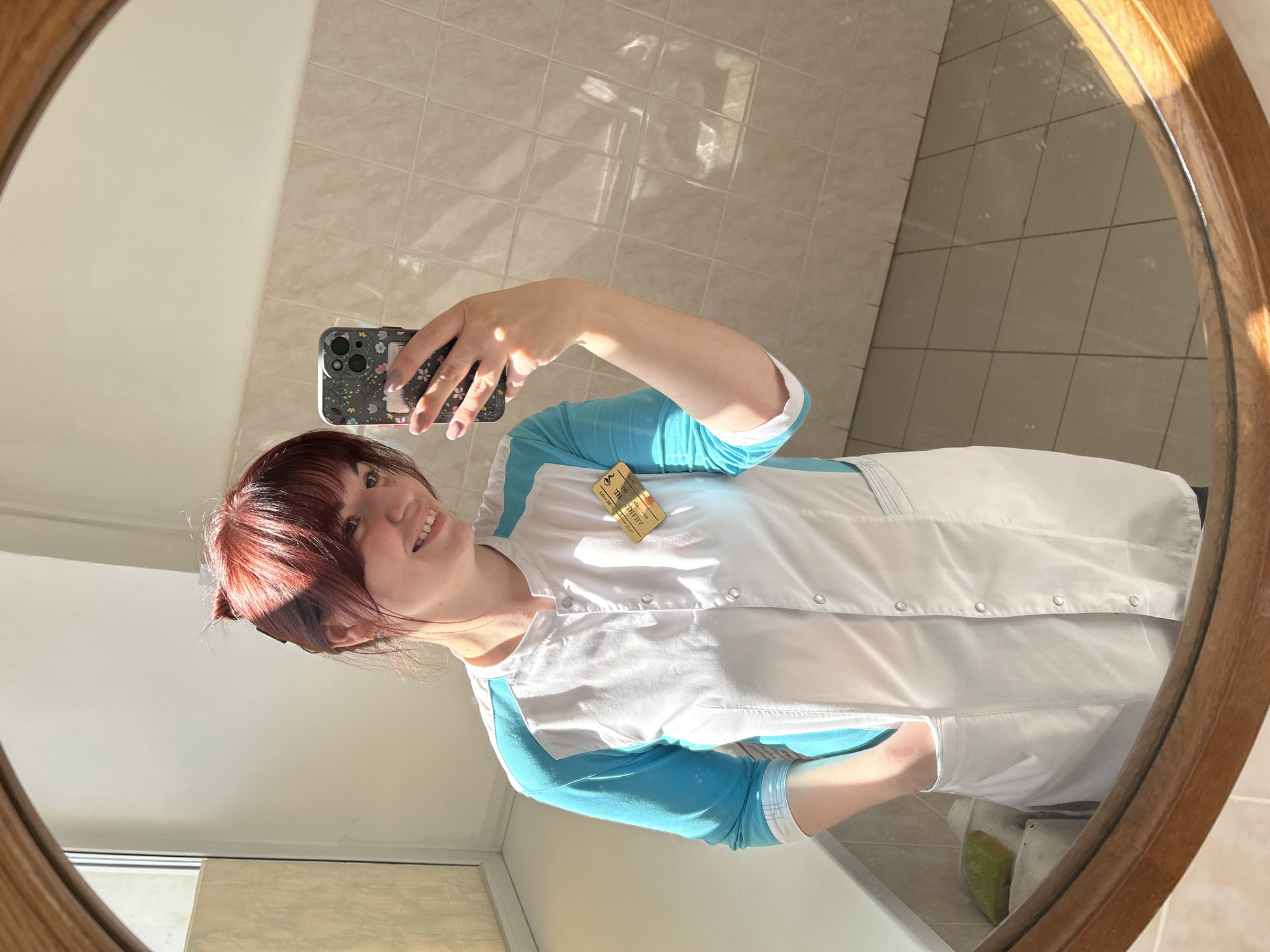 A woman is taking a selfie in a bathroom mirror. She has red hair, and is wearing a white medical coat over a blue long-sleeved shirt, with a name tag pinned to her chest. She is holding a smartphone with a patterned case. The bathroom has light beige tiled walls. The photo appears to be taken indoors, with a window visible on the left side. Based on the name tag, the location is Фоки, Russia.