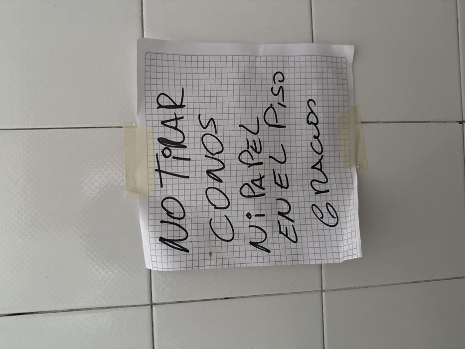 A handwritten sign in Spanish is taped to a tiled wall. The sign reads "NO TIRAR CONOS NI PAPEL EN EL PISO GRACIAS," which translates to "Do not throw cones or paper on the floor, thank you." The sign is written on a piece of graph paper using black ink. Two strips of tape, one on the top-left and one on the top-right, secure the sign to the white tiled surface.