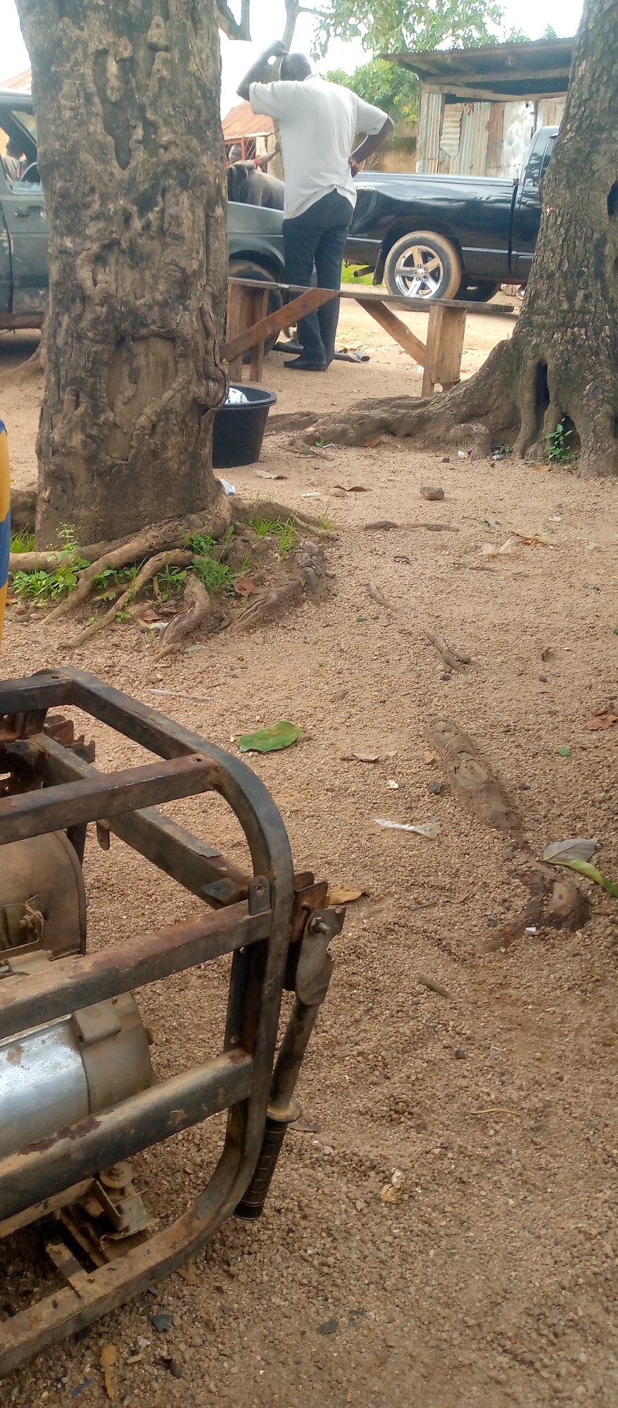 The image shows an outdoor scene with a person standing next to a black pickup truck. The person, viewed from the back, is wearing a light-colored shirt and dark pants, with one hand on their head. The pickup truck appears to be parked on a dirt or gravel surface, surrounded by trees and some rudimentary structures. In the foreground, a rusty metal frame of what appears to be a generator is visible. The environment is sandy with scattered debris, including leaves and some trash. The lighting suggests it is daytime, possibly overcast.