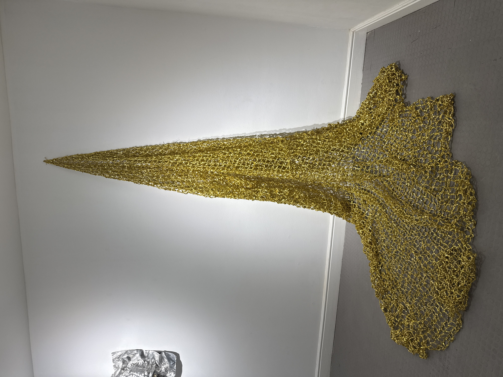 The image shows a piece of art displayed in a gallery. The art appears to be a golden net-like structure, draped from the white wall onto the grey floor. The structure narrows to a point at the top and widens as it extends onto the floor. In the bottom left corner, there's another piece of art, possibly made of crumpled aluminum foil. The location appears to be inside a modern art gallery with clean, minimalist walls and floors. The city and specific location within the city are unknown.
