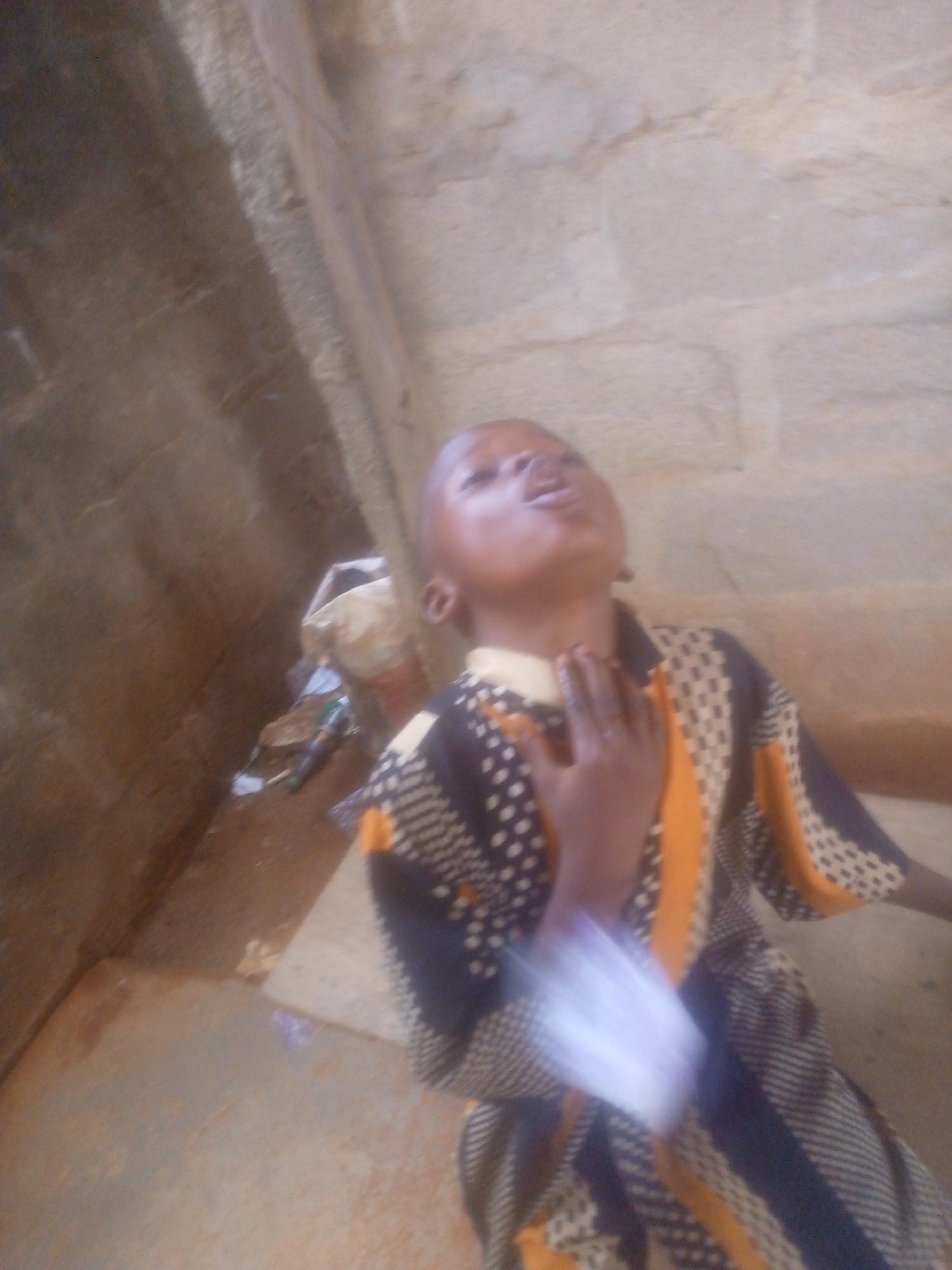 A child stands near a wall in an unspecified location in the city of Unknown, Unknown. The child is wearing a patterned garment and has their hand at their throat. A pile of miscellaneous items sits nearby on a step. The area is surrounded by concrete structures.