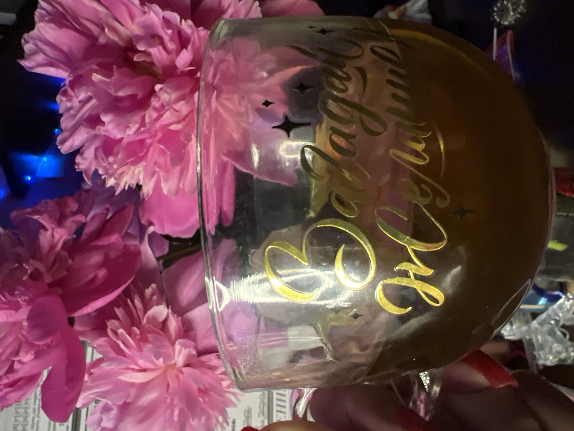 A close-up shot captures a glass with a dark liquid, possibly tea or coffee, sitting in front of a bouquet of pink peonies. The glass is adorned with golden, cursive text that appears to read "Загадай желание," which translates to "Make a wish" in Russian. Several small, dark star-shaped accents are also visible on the glass. A hand with red-painted fingernails is partially visible on the left side of the frame, holding the glass. The background is softly blurred, revealing hints of blue lights and other objects, suggesting an indoor setting. There is no discernible information within the image to indicate the specific location in Minsk, Belarus.