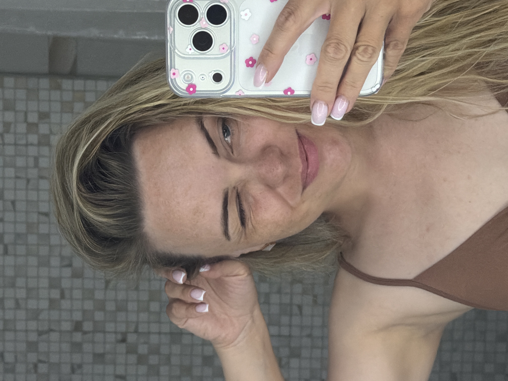 A woman with blond hair, wearing a brown top, is taking a selfie in what appears to be a bathroom, based on the tiled background. She is holding a white smartphone with a transparent case decorated with pink flowers in her right hand. Her nails are manicured with a French tip style. The woman is winking with her left eye and touching her hair with her left hand. The location is not discernible and cannot be identified as Votkinsk, Russia.