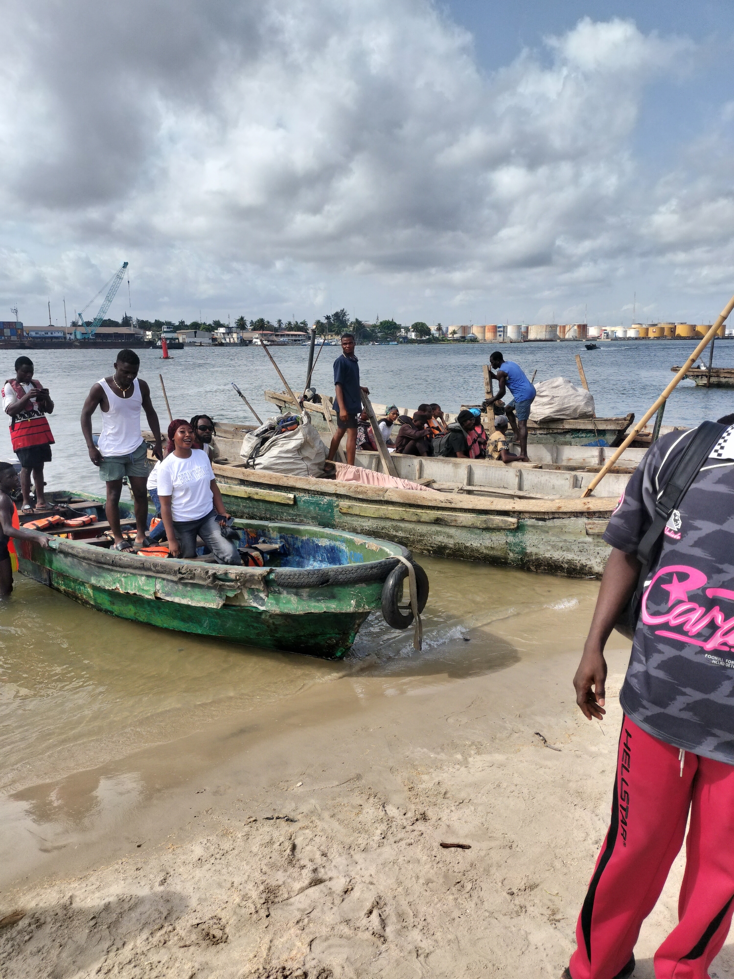 The scene captures a gathering of people around two small boats on a beach, potentially within the Lagos, Nigeria area due to the presence of multiple storage tanks or large industrial containers visible across the water in the background. The boats appear to be constructed from wood, with one boat colored in green and blue details, while the other has a more worn, aged appearance with lighter tones. People are gathered in and around the boats, some standing on the beach. One individual is seen wearing red sweatpants and a gray t-shirt is partially in the foreground and obscures the full view of the boats and people. The weather looks overcast, with a cloudy sky providing diffused lighting on the scene. In the distance, a crane and buildings suggest a port or industrial area.
