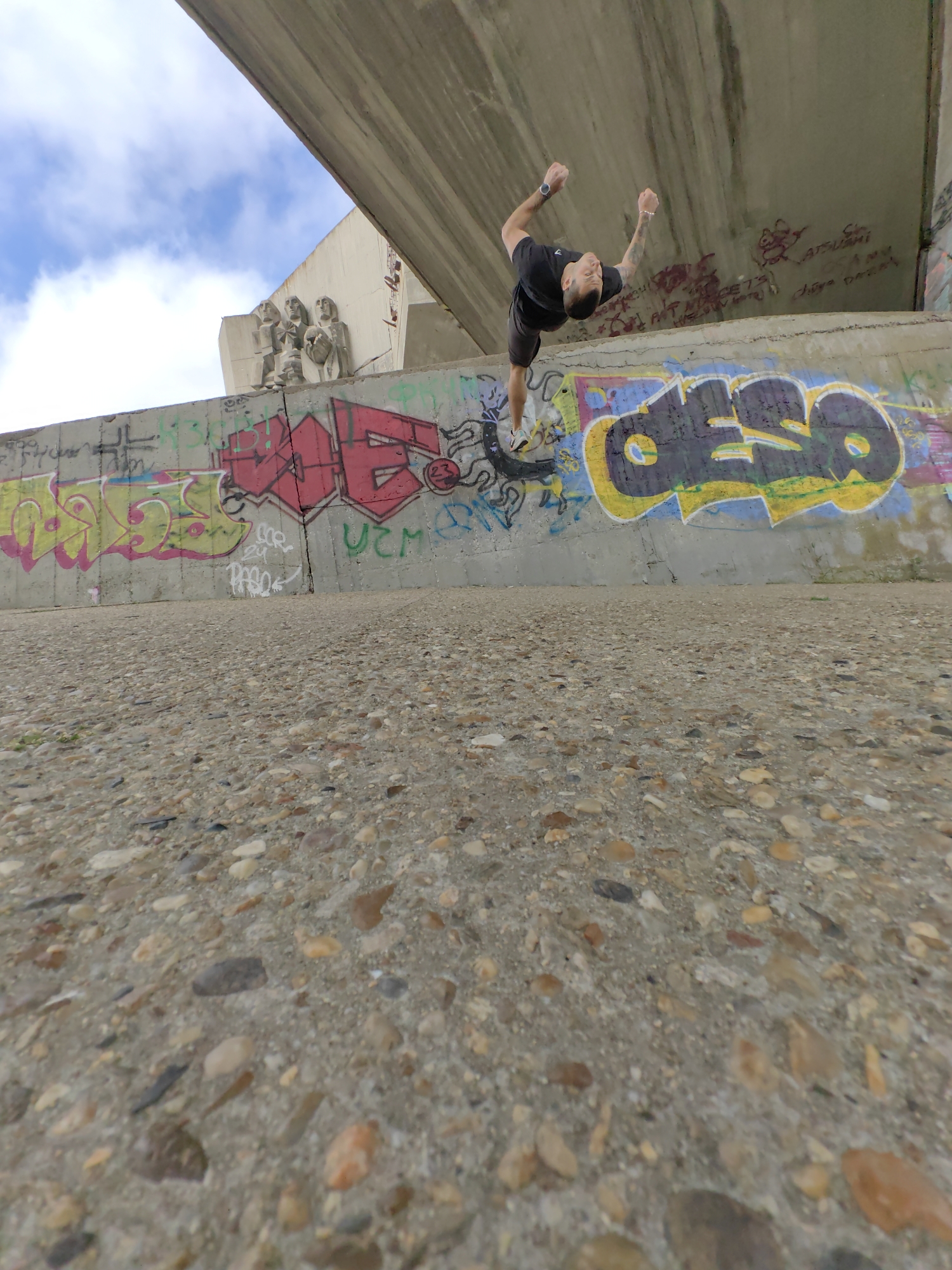 A low-angle, wide-lens photograph captures a man in the midst of a backflip. He is positioned between the viewer and a concrete wall covered in graffiti. The graffiti features lettering in red, yellow, and other colors. Above the wall, partially obscured by a concrete structure, are sculptures of figures. The sky, visible in the upper-left corner, is blue with clouds. The ground in the foreground is paved with small stones embedded in concrete.
Based on the available details (concrete construction, sculptures, graffiti, and style of architecture), the location appears to be near the Monument of the Bulgarian-Soviet Friendship in Varna, Bulgaria.