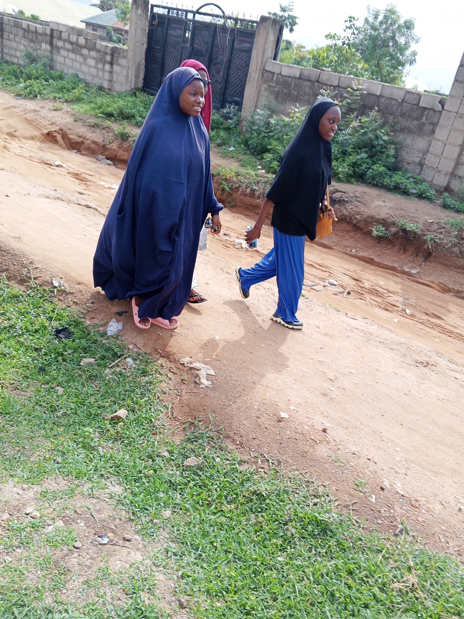 Two people wearing hijabs walk on a dirt road alongside a grassy area in the city of Unknown, Unknown. One person is wearing a long, dark blue garment and pink sandals. The other person is wearing a black hijab, a black shirt, blue pants, and blue sneakers, and carrying a small brown bag. There are brick walls and gates in the background.