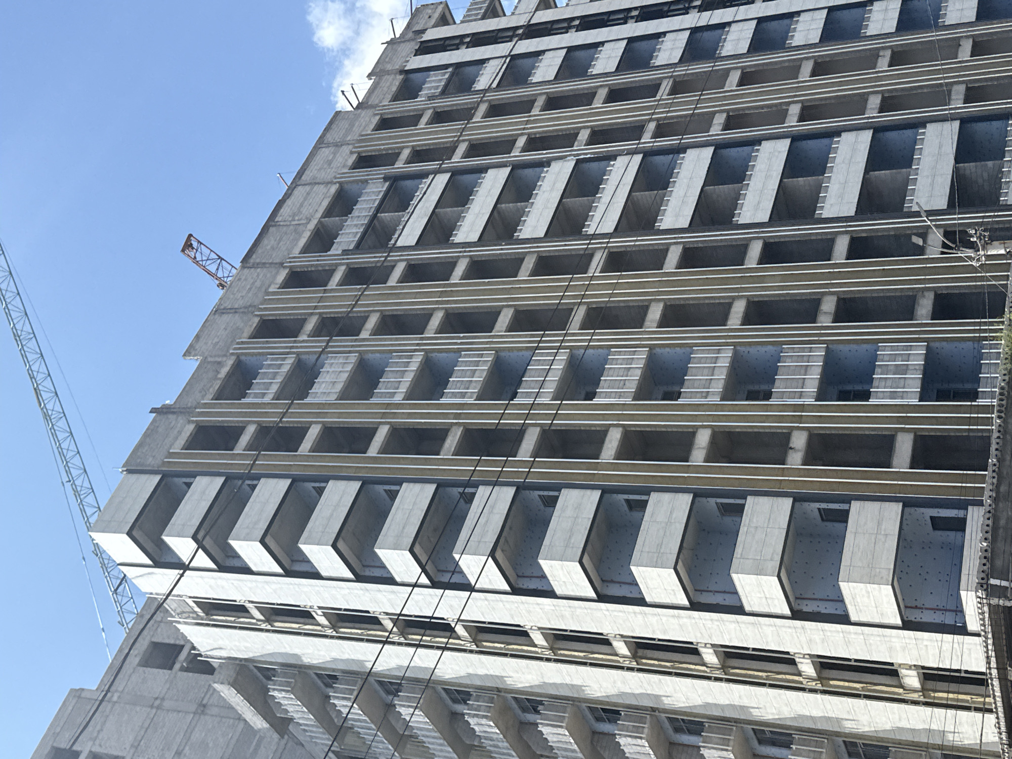 A low-angle shot captures a tall, unfinished building in Yerevan, Armenia, under a partly cloudy, pale blue sky. The building is constructed from grey concrete and features a repetitive pattern of recessed windows and protruding rectangular balconies. A large construction crane stands to the left of the building. Power lines run along the facade of the building.