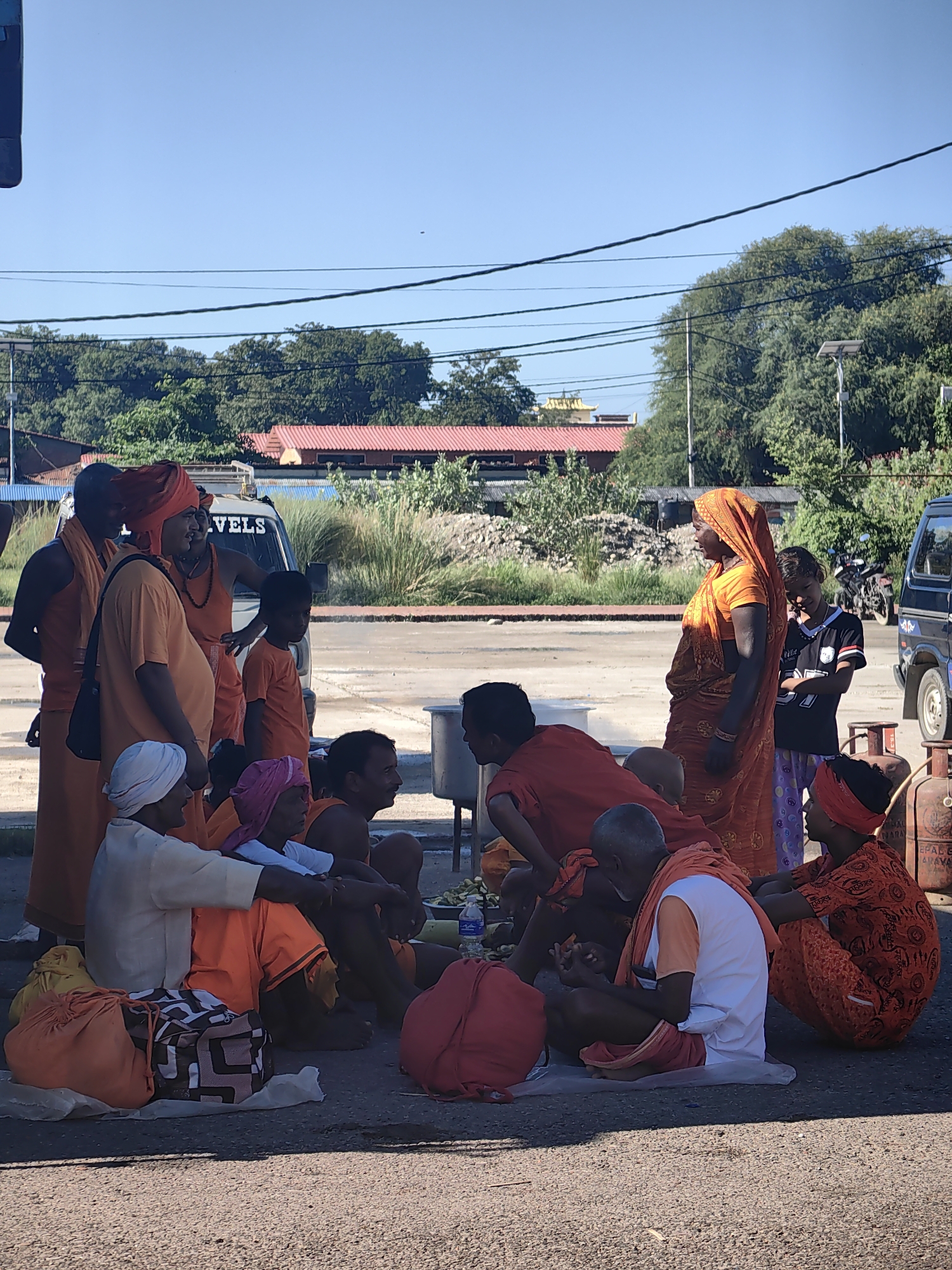 A group of people, mostly men dressed in orange and saffron robes, are gathered on the ground in what appears to be an outdoor public space. Some are sitting on the ground, while others are standing. There is a white van in the background with the word "VELS" visible on its side. A woman in an orange sari and a young boy in a black t-shirt and patterned pants are also present. In the background, there are trees, buildings with red roofs, and what appears to be a temple with a golden roof. There are also gas cylinders and bags on the ground. The scene is brightly lit by sunlight, suggesting it is daytime. The overall impression is one of a gathering or community event taking place in an urban or semi-urban environment in Parsa, Nepal.
