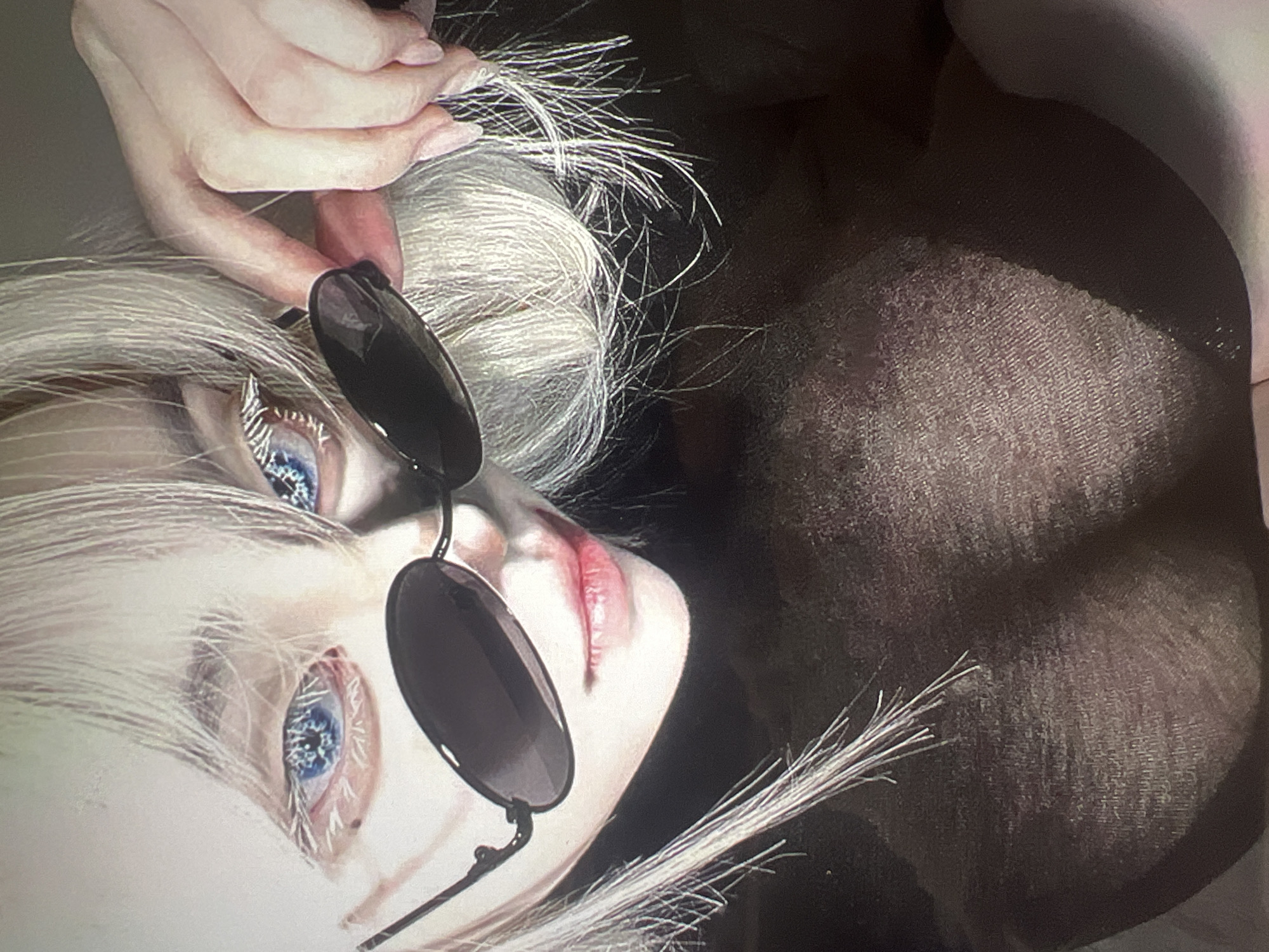 The image is a close-up of a person wearing dark sunglasses and a dark, sheer top. The person has platinum blonde hair, and their eyes, visible through the lenses, are bright blue. Their right hand is adjusting the sunglasses. The background is dark and out of focus. There is no information to indicate the location is Minsk, Belarus.