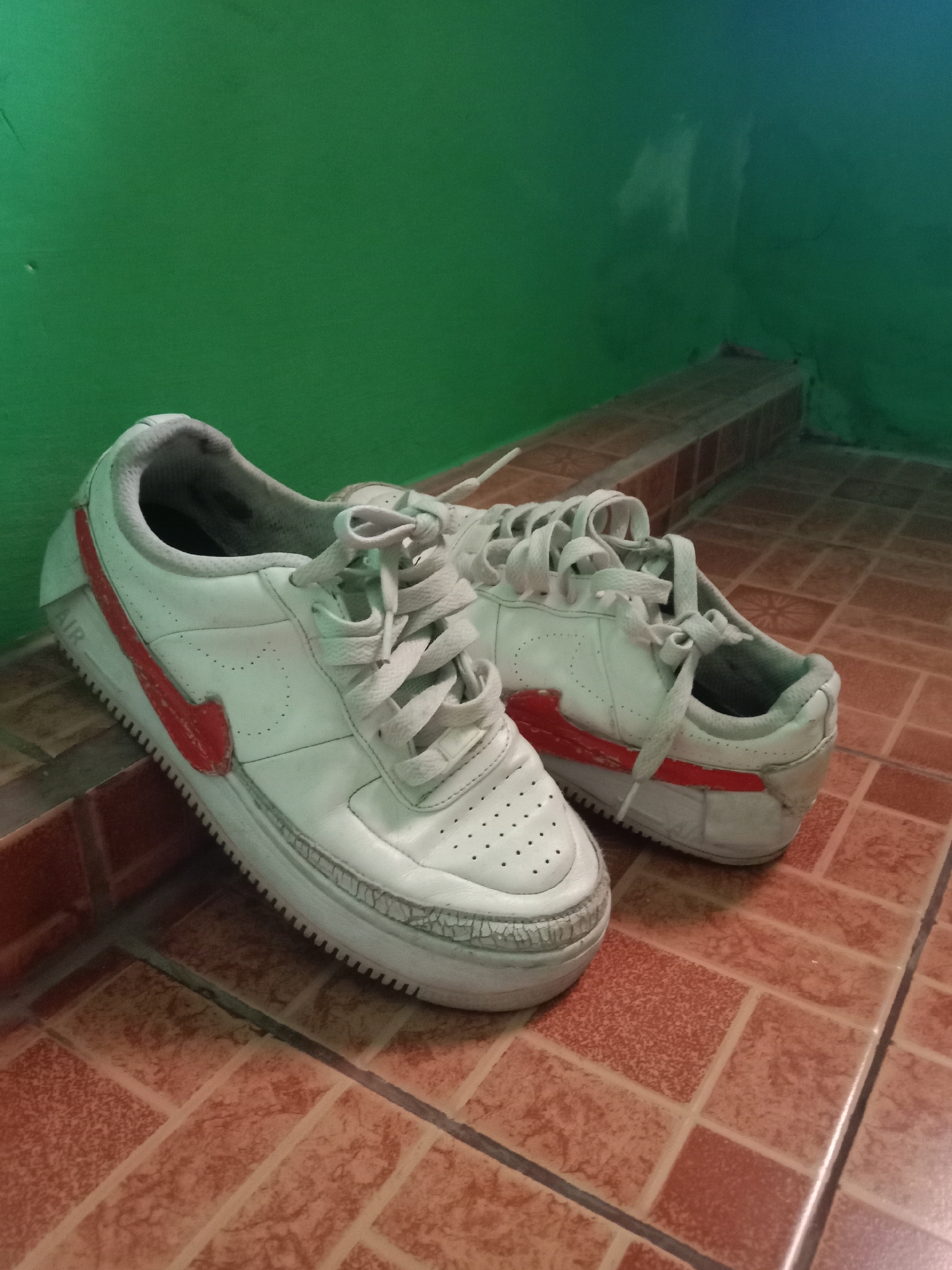 The image shows a pair of white Nike Air Force 1 sneakers with red accents, resting on a terracotta tiled floor. The sneakers appear worn and slightly dirty. They are positioned on a raised ledge or step, with a green wall in the background. The lighting is dim, casting shadows on the floor and wall. The scene is indoors, likely in a residential setting in Kutosari, Indonesia, as indicated by the architectural style and materials often found in the region. No people are present in the image.
