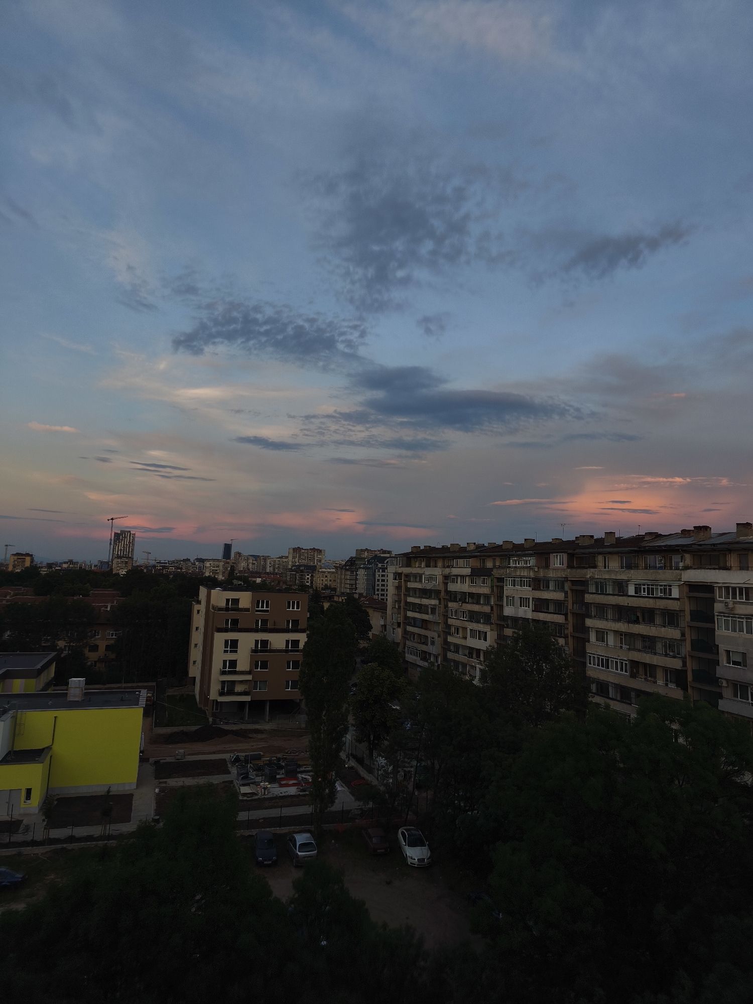 The image captures a city skyline at dusk in Sofia, Bulgaria. The sky displays a gradient of colors, transitioning from a pale blue at the top to hues of pink and orange near the horizon, with scattered clouds. Dominating the foreground are several apartment buildings, characteristic of Soviet-era architecture, with their repetitive balconies and utilitarian design. To the left, a brightly colored yellow building stands out, suggesting a modern construction or renovation. Trees are interspersed throughout the scene, casting long shadows and adding a natural element to the urban landscape. The overall impression is one of a typical residential area in Sofia during the evening.