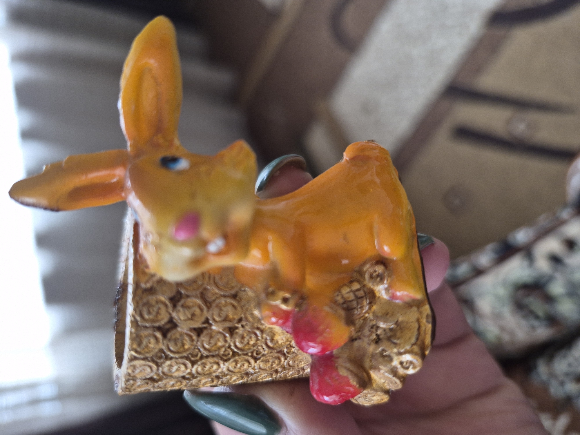 An orange, ceramic figurine of a rabbit sitting on a decorative, gold-colored base is held in a person's hand. The rabbit has blue eyes and pink cheeks, and it is holding two red objects in its paws. The base is cylindrical and textured with a pattern of interwoven circles. The background is blurred and appears to be an indoor setting with a window and curtains. The image was taken on March 20, 2023, in Vanadzor, Armenia.