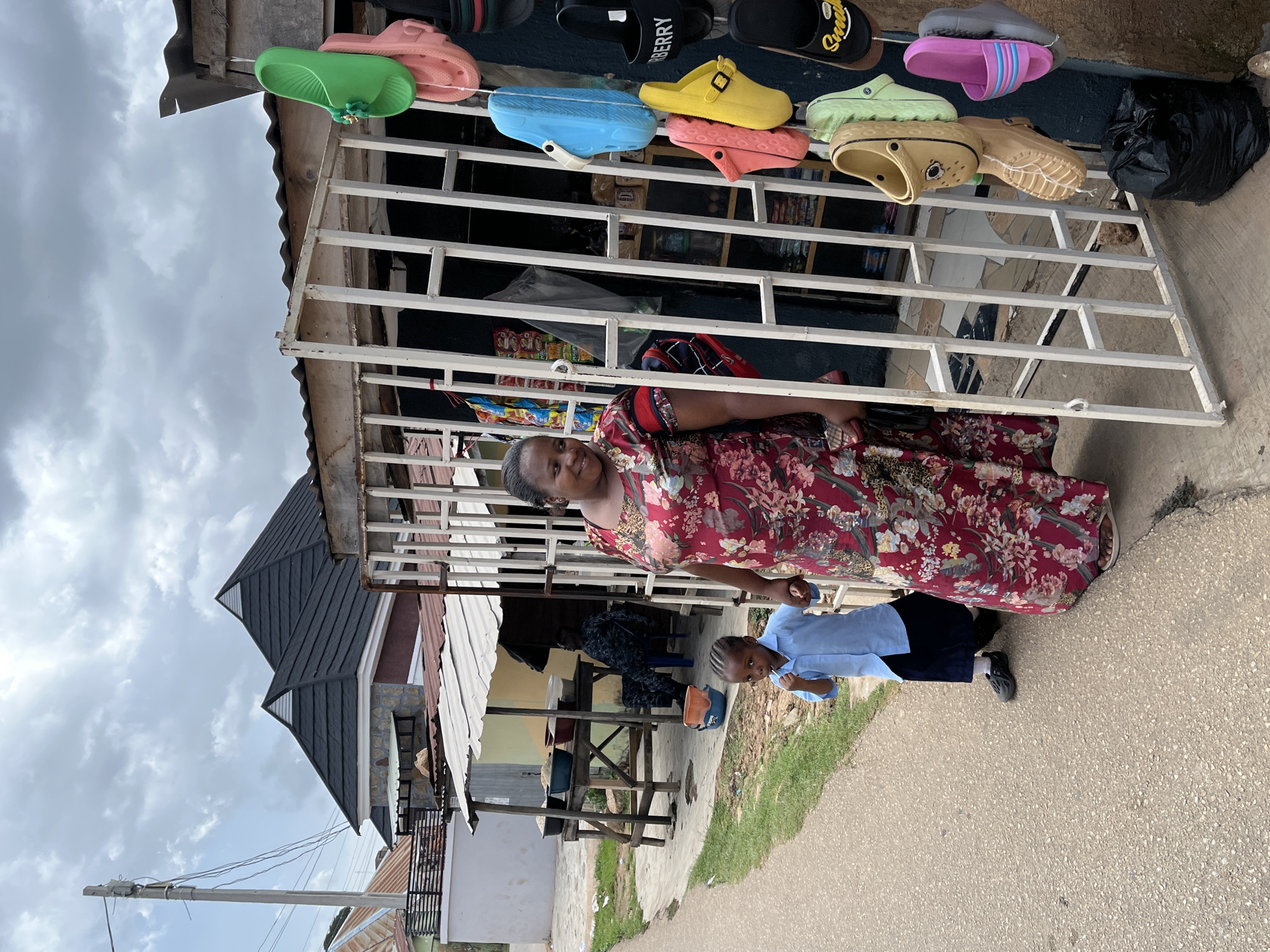 The image, taken in Jos, Nigeria, features a woman and a child standing in front of an outdoor market stall on an overcast day. The woman, with dark skin and dark hair, wears a long-sleeved, red dress with a prominent floral pattern. She is smiling and looking directly at the camera, holding the hand of a young child beside her.
The child, also with dark skin, is dressed in a light blue long-sleeved shirt and dark trousers, appearing to be a school uniform, and is wearing dark slip-on shoes. The child is looking slightly away from the camera.
Behind the woman and child is a market stall constructed with a white metal grate framework. Numerous colorful clogs and slippers in various hues like green, pink, black, blue, yellow, light green, beige, grey, and purple are displayed on these grates. Visible text on some of the black slippers includes "BURBERRY" and "SNEAKERS." Inside the stall, packaged goods are faintly visible.
In the background, other buildings with corrugated metal roofs and walls are present, including one with a distinctive black-tiled, multi-pitched roof. A utility pole with overhead wires is visible on the left. The ground surface is a mix of concrete, dirt, and some grass. The scene captures a daily life moment in a local market setting.