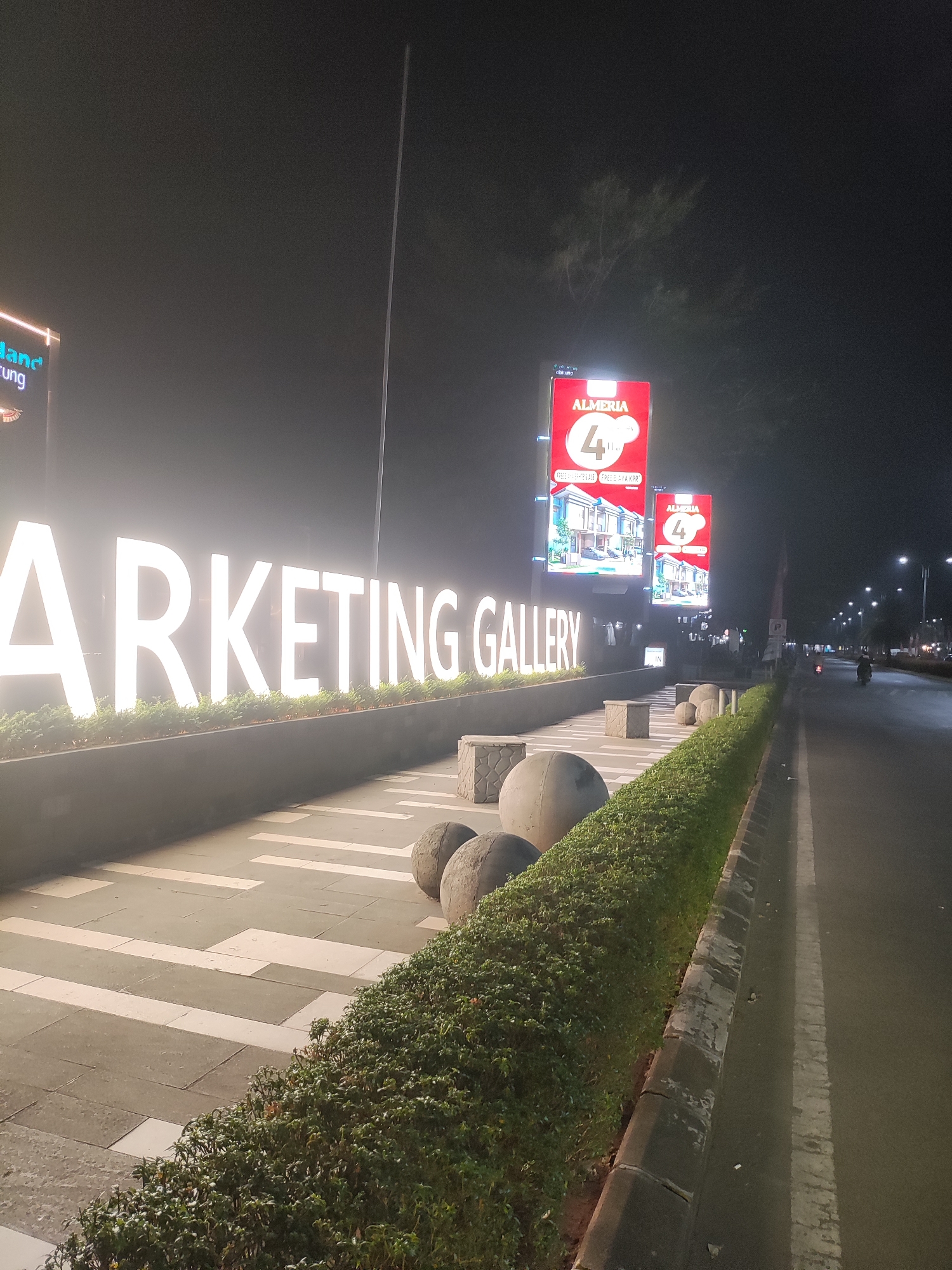 The image depicts a night scene on a street in Telagaasih, Indonesia, illuminated by artificial lights. A large, brightly lit sign spelling out "MARKETING GALLERY" is prominently displayed on the left side of the frame. The letters are white and stand out against the dark background. Behind the sign, there are tall, vertical billboards featuring advertisements for "ALMERIA" and a "4th" anniversary promotion with images of houses.
In the foreground, a paved walkway with a geometric pattern of light and dark tiles lines the "MARKETING GALLERY" sign. A neatly trimmed green hedge runs along the edge of the walkway, separating it from the road. Several large, spherical stone sculptures are placed near the hedge and walkway.
To the right of the walkway and hedge is a paved road with a white lane marking. The road is dimly lit by streetlights, and a few vehicles, including a motorcycle with a rider, are visible in the distance. The overall atmosphere is urban and commercial, characteristic of a developing area in Indonesia at night. The sky is dark and hazy, suggesting artificial lighting and possibly some atmospheric moisture.