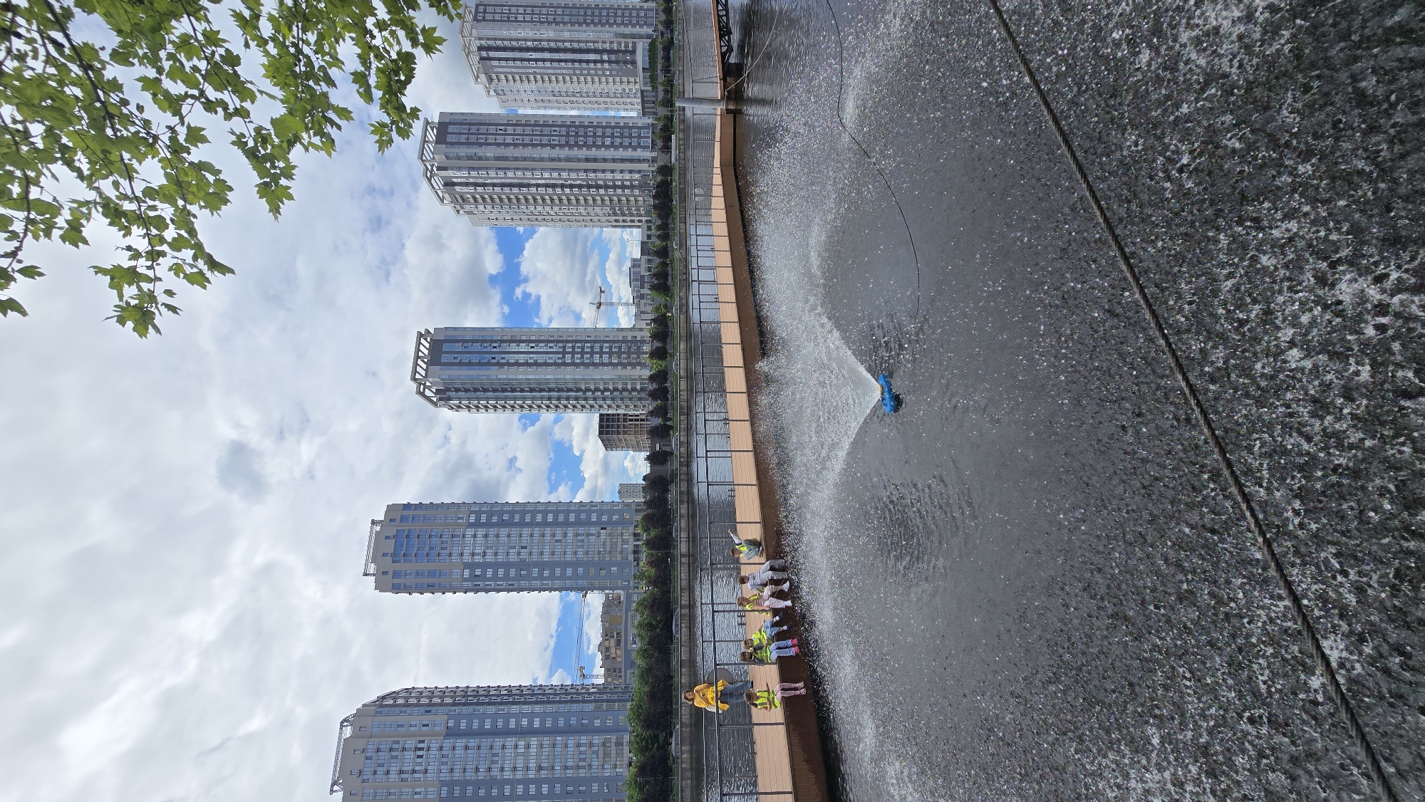 The image depicts a waterfront scene in Kyiv, Ukraine, with a row of modern high-rise apartment buildings forming the backdrop. In the foreground, a wooden pier extends into a body of water where a blue sprinkler is spraying water. Several children, wearing high-visibility vests, are seated on the pier, some looking towards the water. An adult, also in a yellow high-visibility vest, stands near the edge of the pier. The sky is overcast with scattered clouds. Trees line the shore behind the pier.