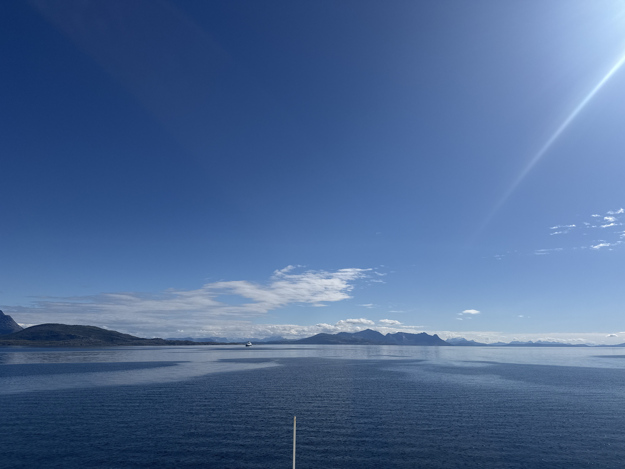 A wide-angle, high-angle shot captures a serene seascape under a bright, clear blue sky with a prominent sunbeam extending from the upper right corner. The foreground is dominated by the deep blue, rippling surface of the water, with a single white pole extending vertically from the bottom center. In the mid-ground, a distant white ferry is visible sailing across the water. The background features a chain of majestic, dark, and somewhat rugged mountains, some with snow-capped peaks, receding into the distance. The coastline is dotted with islands and fjords, typical of the Norwegian landscape. Wispy white clouds are scattered across the sky, adding depth and texture to the scene. The overall impression is one of tranquility and natural beauty. The scene is consistent with views from the town of Lødingen, Norway, known for its stunning fjord and mountain scenery.