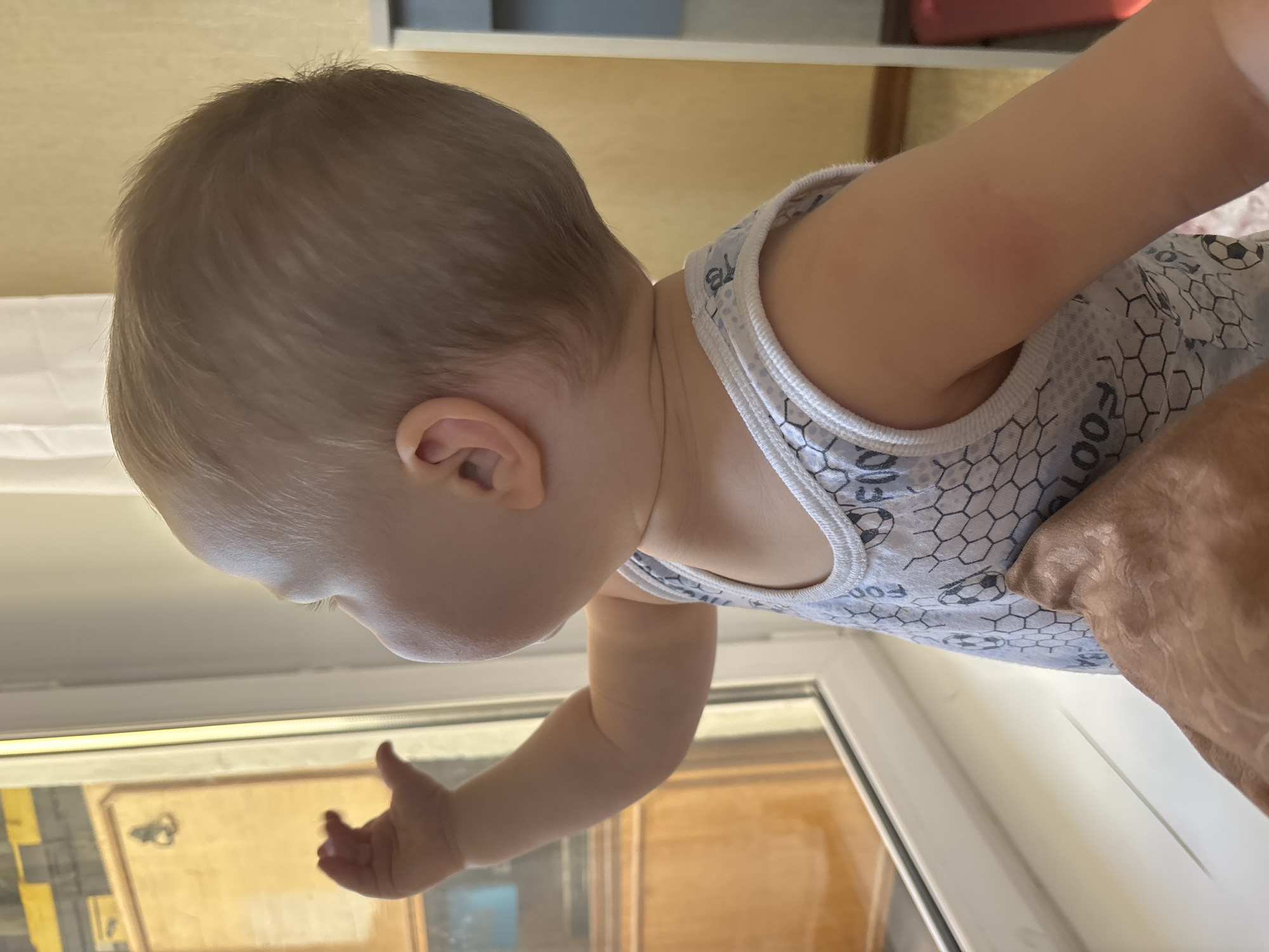 A young child, with light brown hair and fair skin, stands facing a window. The child wears a white sleeveless shirt with a pattern of soccer balls and hexagonal shapes. The child's right arm is extended towards the window, with the hand open and fingers slightly curled, as if reaching for something. The child's head is turned to the left, with the profile visible, and the child is looking out of the window. A portion of an adult's arm, covered in a light brown garment, is visible in the lower left corner of the image, positioned near the child's torso. The windowpane is clear, revealing a blurred interior scene with wooden elements and a framed object. The environment appears to be indoors, with a yellow wall in the background and a white curtain visible on the right. The window frame is white, and the windowsill is also white. The scene suggests a domestic setting, possibly an apartment in Naberezhnye Chelny, Russia, given the prompt's inclusion of this location.