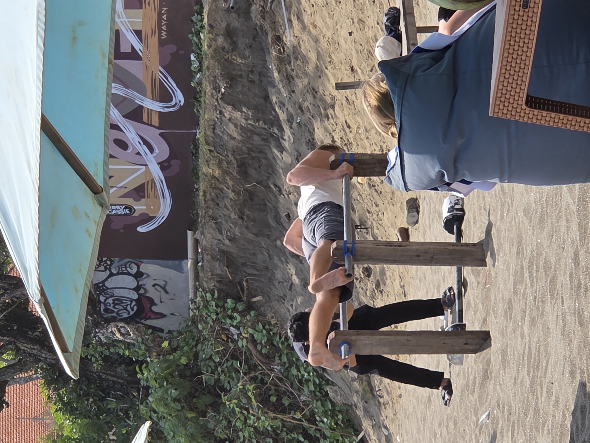 A beach scene in North Kuta, Indonesia, features two individuals on a makeshift wooden structure. One person, wearing shorts and a white shirt, is positioned higher, gripping the wood. The other, in black pants and a gray shirt, is below, also holding the wood. The structure appears to be set on sand. In the background, there's a mural wall with the word "WAYAN." An umbrella is partly visible on the left side. Other details include greenery and what seems to be the edge of a building.