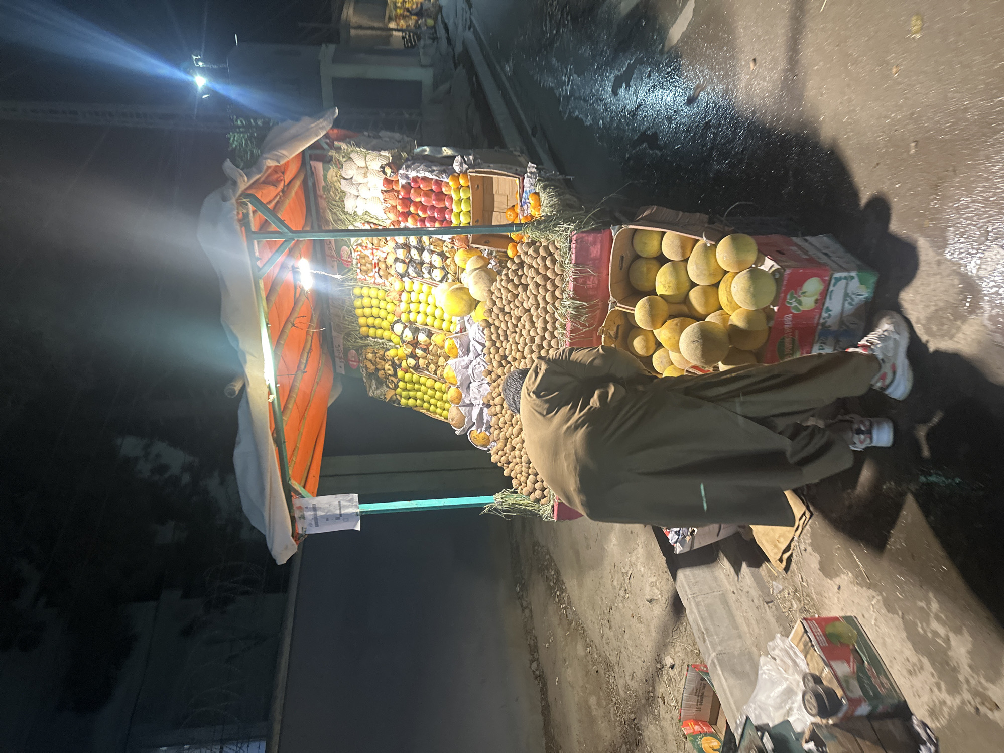 A nighttime street scene in Quetta City Tehsil, Pakistan, shows a fruit stand brightly illuminated by artificial light. The stand displays a variety of fruits, including apples, oranges, mangoes, and longans, arranged in neat piles. A person wearing a brown robe stands partially obscured in front of the stand. The ground around the stand is wet and littered with debris. Streetlights provide additional illumination in the background.