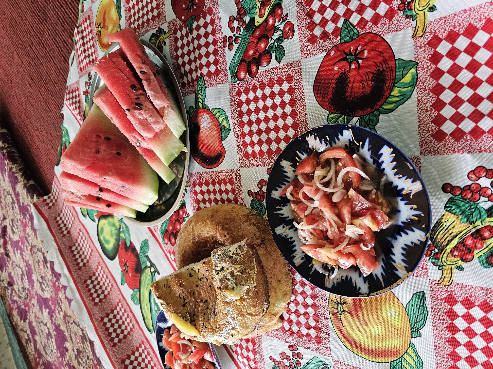 A close-up, overhead shot captures a spread of food laid out on a table covered with a patterned tablecloth. The tablecloth features a checkered red and white design interspersed with illustrations of fruits and vegetables, including tomatoes, apples, and cherries.
In the center of the frame is a large, round loaf of bread, split open to reveal a cheesy, golden-brown interior. To the left of the bread, a small, dark blue and white patterned bowl holds a salad of chopped tomatoes and thinly sliced onions. To the right of the bread, a metal platter is filled with several slices of watermelon, some cut into wedges and others into sticks. The watermelon slices are vibrant pink with dark seeds. Another small, dark blue and white patterned bowl containing a similar tomato and onion salad is partially visible in the lower left corner of the image.
The scene is likely a meal setting, possibly in Özgön, Kyrgyzstan, given the style of the bread and the common presence of these foods in the region's cuisine. The lighting suggests an indoor setting with natural light or overhead illumination. No people are visible in the image.