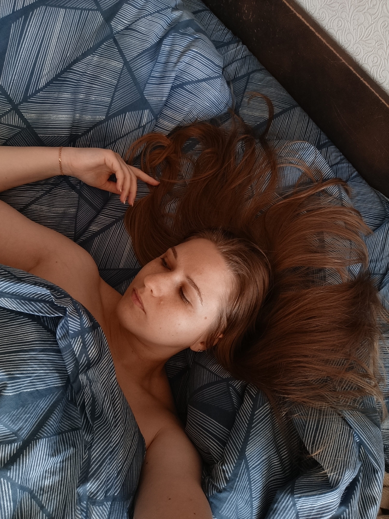 A young woman with long auburn hair lies on a bed covered with a blue and white striped duvet. Her eyes are closed, and her head is tilted slightly to the side. Her right arm is bent at the elbow, with her hand resting on the duvet. She is wearing a bracelet on her left wrist. The background is a dark brown headboard and a patterned wallpaper. The scene appears to be indoors, in a bedroom.