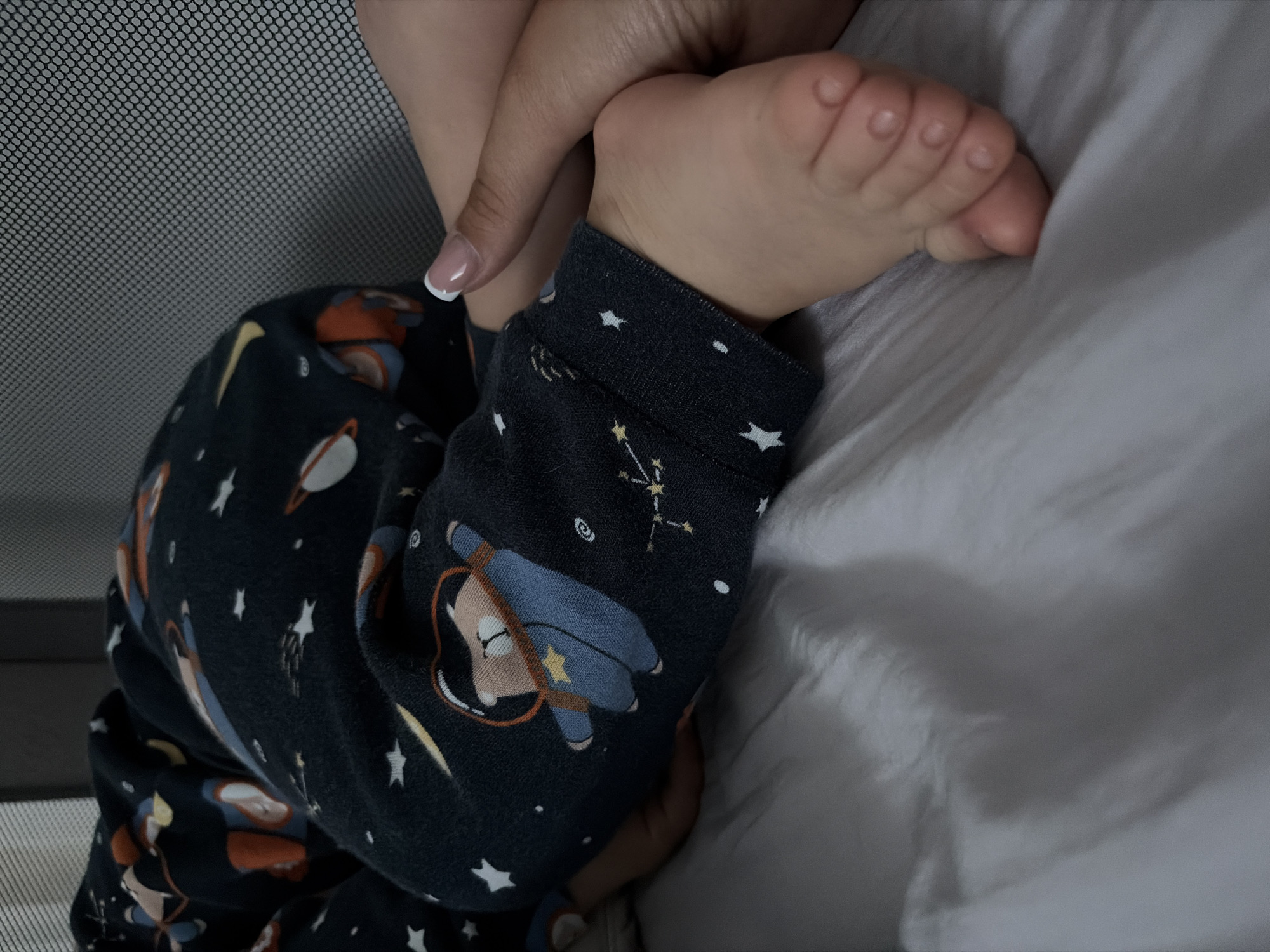 The image captures a tender moment featuring a baby's foot and leg, gently touched by an adult's hand. The setting appears to be indoors, likely a bedroom or nursery, with a soft, muted light, suggesting evening, night, or early morning.
The baby's leg is clad in dark-colored pajamas adorned with a playful space theme. Visible patterns include multiple white stars, a constellation diagram, and cartoon astronauts, one with a prominent yellow star on its helmet, as well as planetary figures resembling Saturn. The fabric of the pajamas appears soft and slightly rumpled.
The adult's hand, with a neatly manicured fingernail featuring a white French tip, delicately holds or rests near the baby's foot. The baby's bare foot, with its small, soft toes, is visible against a white, subtly wrinkled sheet or blanket. To the left, a grey, fine-mesh barrier, characteristic of a crib or playpen, is partially visible. The overall scene conveys a sense of calm, warmth, and affectionate care. No explicit text is visible in the image. The location context provided is Svetovrachene, Bulgaria.