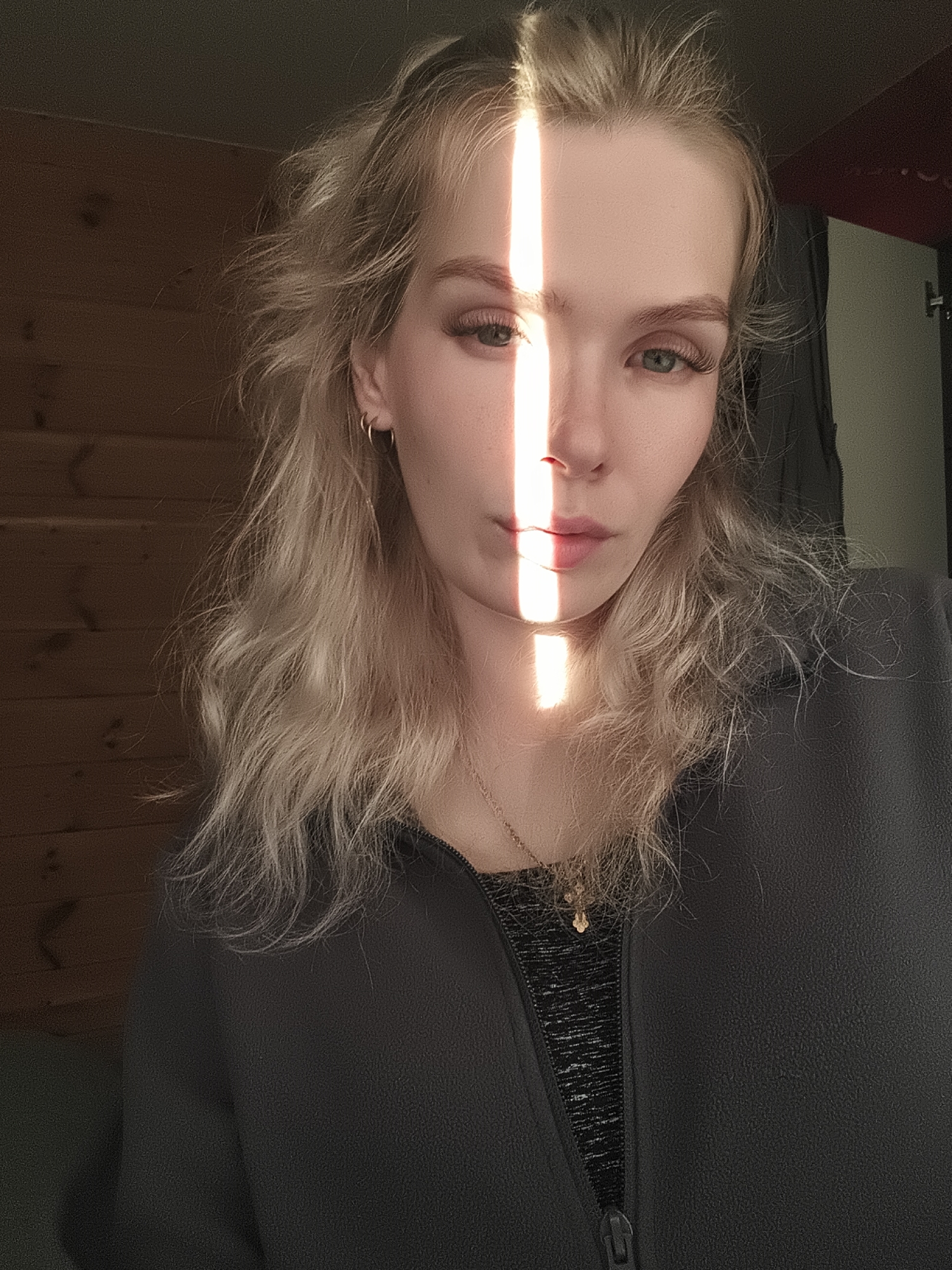This media file is a close-up, medium shot selfie of a young Caucasian woman with blonde, wavy hair. She is looking directly at the camera with a neutral expression. A strong beam of natural light falls vertically across her face, illuminating the right side of her forehead, nose, and upper lip, while leaving the left side in shadow. This creates a dramatic contrast and highlights her green eyes and long eyelashes.
She is wearing a dark gray zip-up jacket over a black and gray patterned top. A delicate gold necklace with a small pendant is visible around her neck. She has a small hoop earring in her left ear.
The background consists of a wooden-paneled wall on the left side of the frame, suggesting an indoor setting with wooden architecture. On the right side, a dark vertical shadow suggests a doorway or a wall with a darker surface. The lighting indicates it is daytime, likely late afternoon or early morning, given the strong, directional sunlight casting distinct shadows. The overall mood is contemplative and slightly dramatic due to the lighting. No text is visible in the image. The location context is Русский Вожой, Russia.