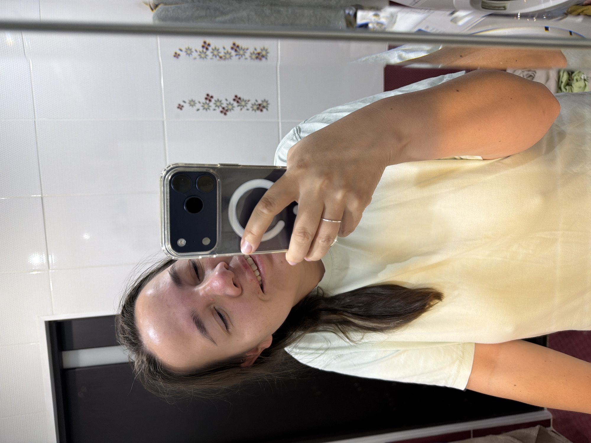 A woman is taking a mirror selfie in a bathroom. She is wearing a light yellow t-shirt. The bathroom has white tiled walls and a dark cabinet. A towel and a washing machine are visible in the background. The woman has her eyes closed and is smiling. Her phone, which is in a clear case, is held up to take the photo. She is wearing a thin silver ring on her right hand.