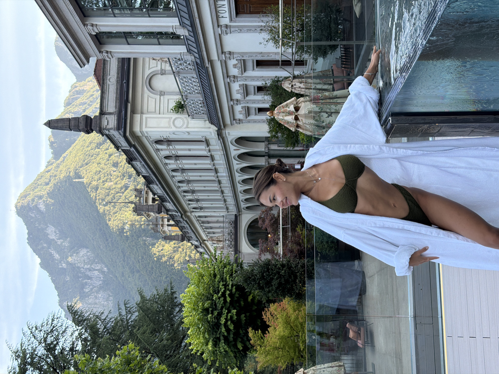 A woman in a green bikini and white robe stands on a deck by a pool with mountains and a historic building in the background. The woman is leaning on the edge of the pool, her body angled towards the left of the frame. Her hair is pulled back into a bun, and she is looking to the left. The building behind her is white with classical architectural elements, including arches and columns. It appears to be a grand, old structure, possibly a hotel or spa. The mountains are covered in trees and have rocky outcrops. The lighting suggests it is late afternoon or early evening. The setting is likely the Grand Hotel San Pellegrino in San Pellegrino Terme, Italy, a historic spa town known for its mineral water and Art Nouveau architecture.