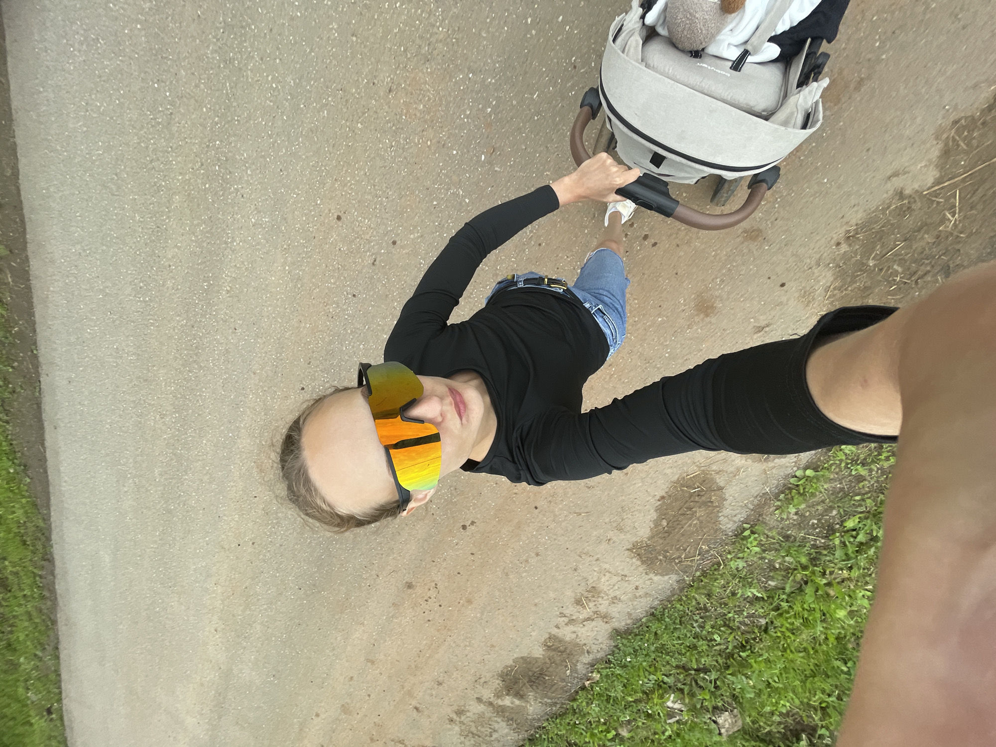 A woman is taking a selfie while pushing a stroller. She is wearing large, reflective sunglasses, a black long-sleeved shirt, and denim shorts. Her right hand is on the stroller handle, which is a neutral beige color with brown grips. The stroller appears to be occupied by an infant, partially visible under a beige cover. The ground is a textured, light-colored surface, possibly asphalt or a dirt path, with some patches of mud and grass on the left side. The lighting suggests it is daytime. The image is taken from a high angle, looking down at the woman and the stroller. The location is identified as Podshivalovo, Russia, though this specific detail cannot be visually confirmed from the image itself.
