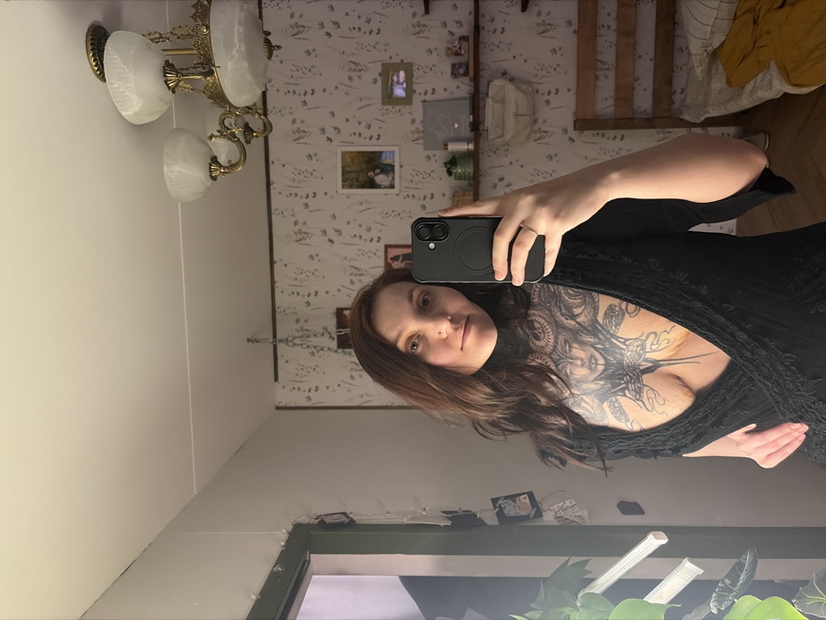 A woman takes a selfie in a mirror. She has brown hair, light skin, and a large tattoo across her chest. She is wearing a low-cut black dress or robe. The background shows a room with patterned wallpaper, pictures on the wall, a chandelier, and a window with plants in front of it. The mirror is slightly angled, capturing part of the ceiling and a wooden structure.