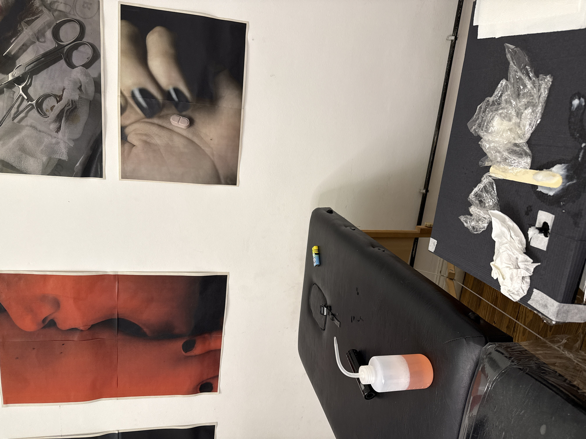 A tattoo artist's workspace is depicted, showcasing various elements associated with the profession. On a white wall, three posters are affixed. The first displays surgical tools on a sterile drape, the second highlights a hand with dark nail polish and a small pill, and the third shows a close-up of faces pressed together in a reddish hue.
To the right, a black tattoo chair is visible with an attached bottle containing a pink liquid. An examination table is arranged with assorted single-use disposable materials and tattoo-related implements.
The location is specified as Buenos Aires, Argentina.