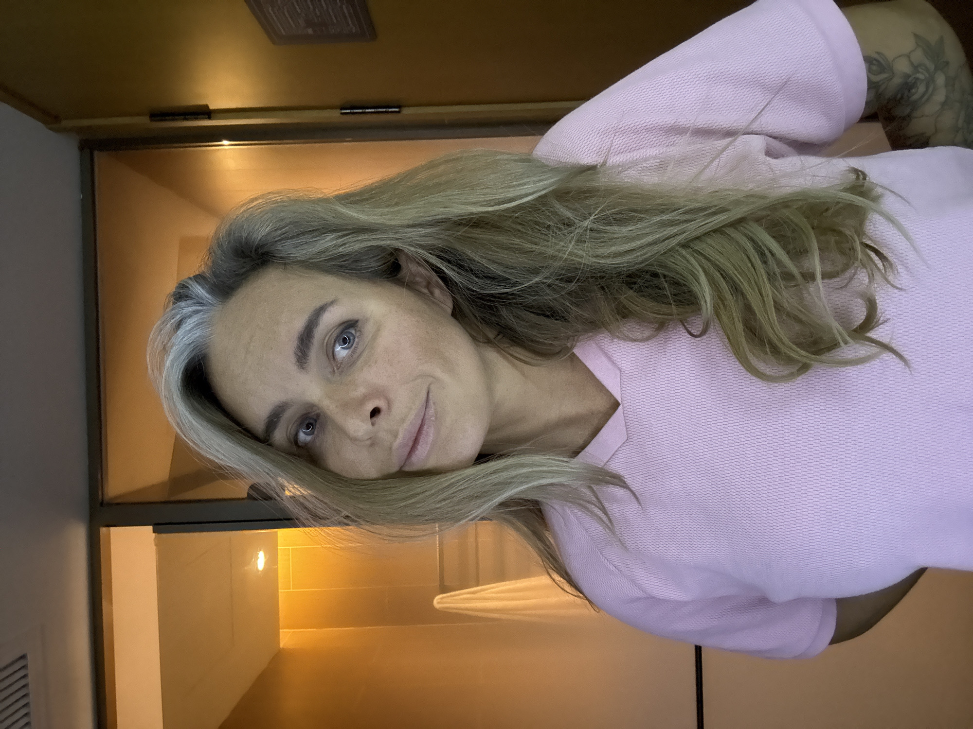 A woman with blonde hair and visible gray roots, wearing a pink collared shirt, is posing for a selfie. She has blue eyes and a slight smile. Behind her, a bathroom with orange-tiled walls and a towel hanging on a rack is visible through a glass partition. A wooden door is on the right side of the image. The scene appears to be indoors, possibly in a hotel room. There is no indication of the location being in Shanghai, China.