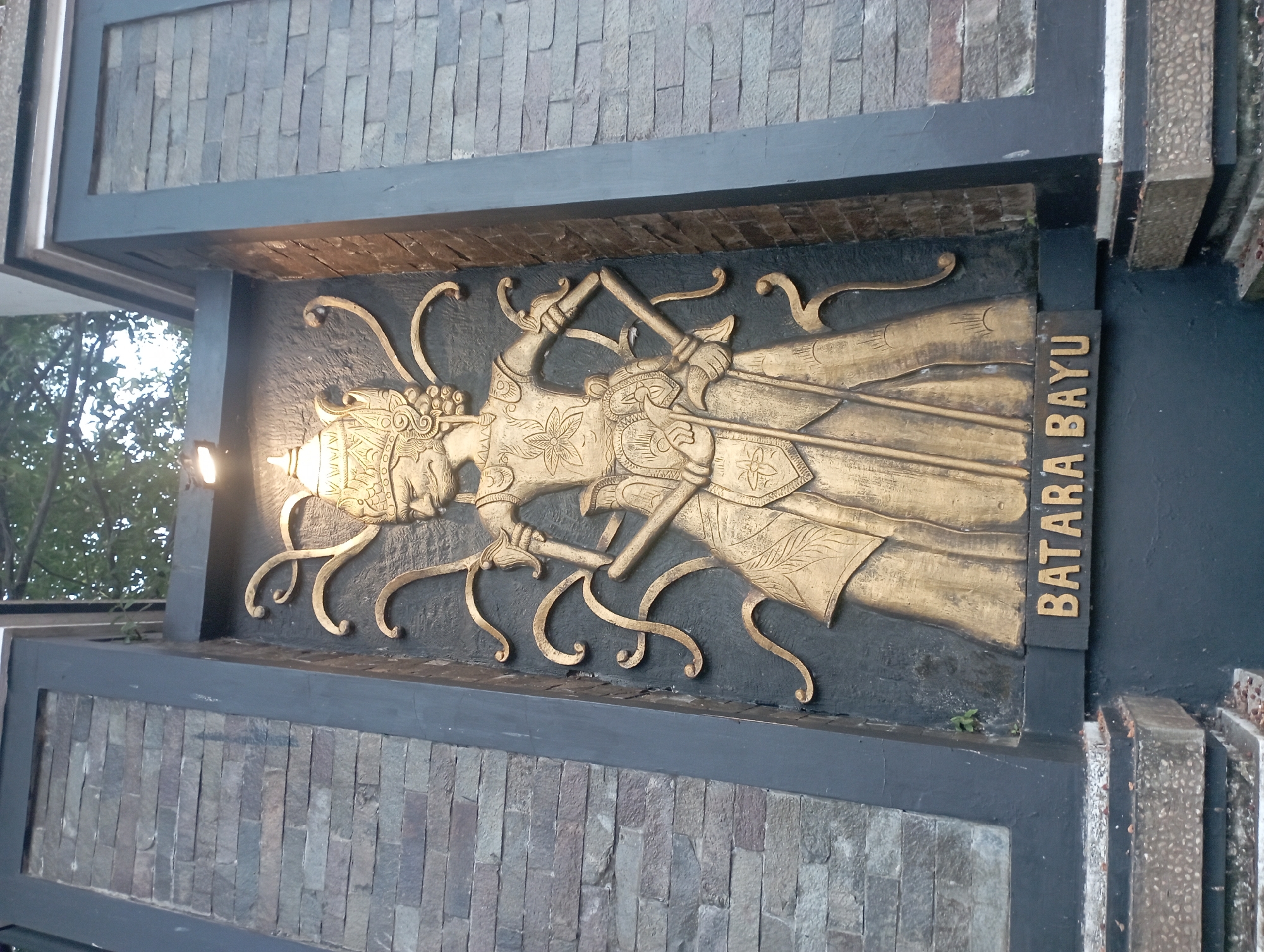 A low-angle, eye-level shot shows a golden relief carving of a figure with elaborate headwear and flowing garments against a dark background. The figure is adorned with intricate patterns and holds a staff-like object. The relief is mounted on a dark grey rectangular panel, which is flanked by a stone-tiled wall on the left and a dark grey pillar on the right. The bottom of the relief has a gold plaque with the inscription "BATARA BAYU". The scene appears to be outdoors, with trees visible at the top and part of a stone structure at the bottom. This relief is located at the entrance to the Sasana Budaya Purwakarta (Purwakarta Cultural Hall) in Purwakarta, Indonesia.