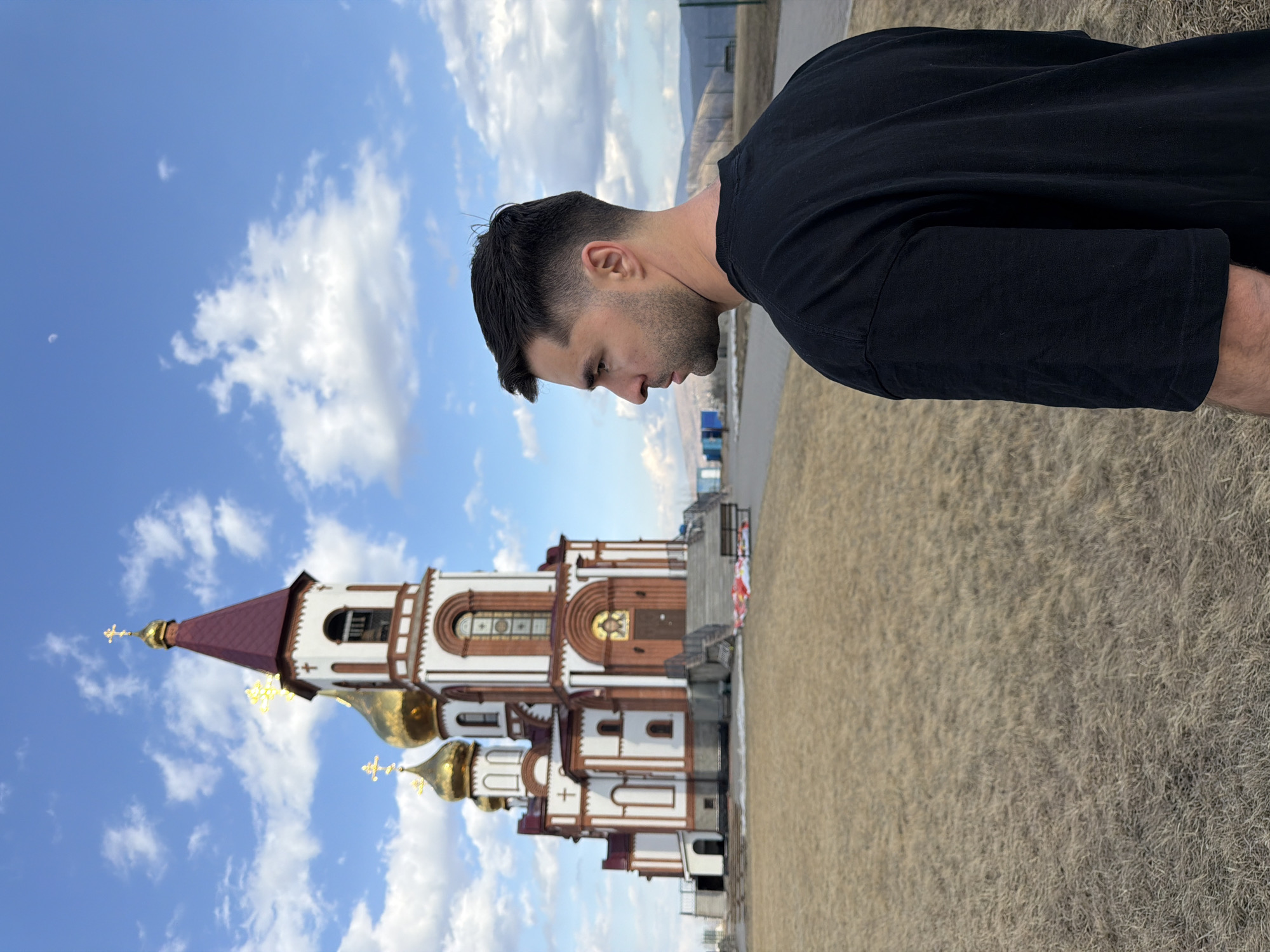The image shows a man standing in front of the Paraskeva Pyatnitsa Chapel in Krasnoyarsk, Russia. The chapel, with its red and gold domes, is positioned in the lower left quadrant of the image and is set against a backdrop of blue sky with white clouds. The man is wearing a black t-shirt and is partially framed on the right side of the image, his gaze directed downwards. The foreground is a dry, grassy area. A road is visible in the background.
