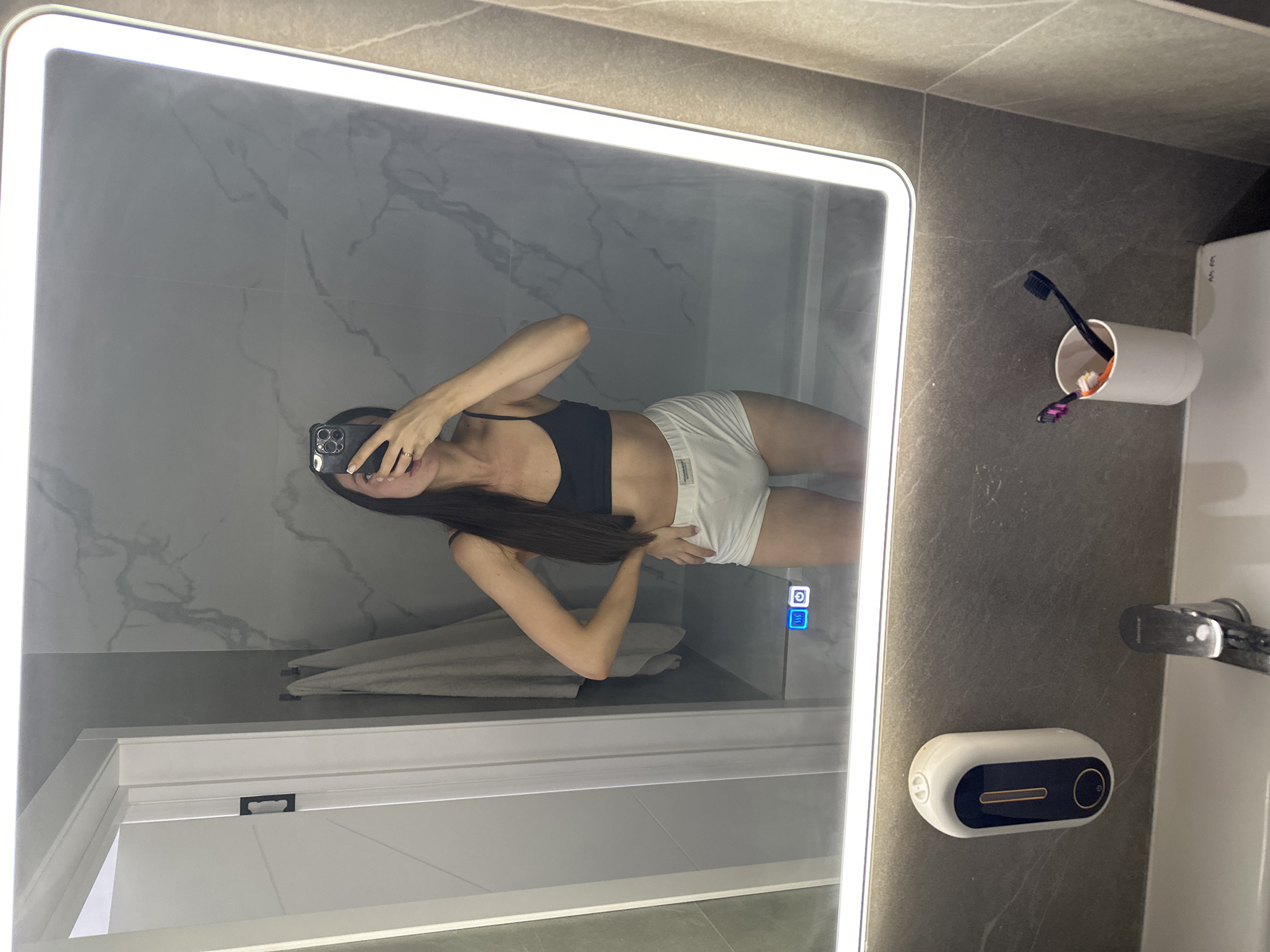 A woman is taking a mirror selfie in a modern bathroom. She is wearing a black sports bra and white high-waisted shorts. Her long, dark hair is down and she is holding a black smartphone with both hands to take the photo. Her left hand is on her hip, and her right arm is bent at the elbow. The bathroom features a large, illuminated mirror with a marble-effect backsplash. To the left of the mirror, a white door with a black handle is visible. Two towels hang on the left side of the image. On the mirror's surface, a blue icon representing heating and a power button icon are visible. Below the mirror, a white soap dispenser is mounted on the wall, and a chrome faucet is situated above a white sink. To the right of the sink, a white cup holds two toothbrushes, one black and one with purple and orange bristles. The overall environment suggests a clean and contemporary residential bathroom. There is no indication of the location being in Эстачи, Russia.