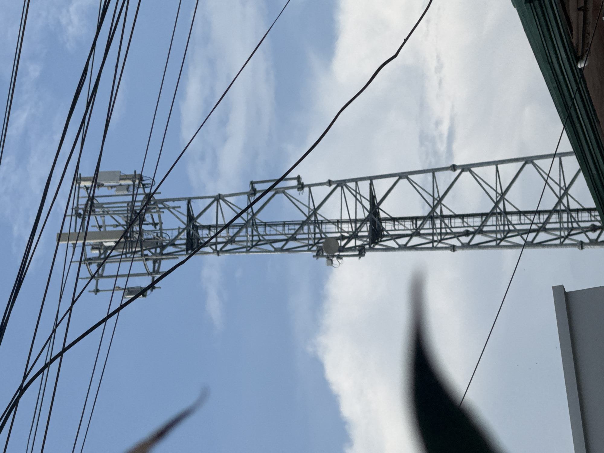 The image, taken in Davao City, Philippines, presents a rotated view of a tall, metallic lattice tower against a partly cloudy daytime sky. The tower, likely a telecommunications or utility mast, stretches horizontally across the middle of the frame, showcasing a complex structure of grey metal beams. Various antenna-like equipment and small boxes are affixed to its framework.
A dense network of dark, slender electrical or utility wires crisscrosses the upper left section of the sky, forming diagonal lines across the frame. The sky itself displays a blend of clear blue patches and soft white clouds.
On the far right, sections of what appears to be a building or architectural structure are visible, characterized by dark green/brown and lighter grey surfaces. A large, blurred, dark shape in the bottom right corner suggests foreground foliage or an object very close to the camera. There is no discernible activity or visible text in the image. The perspective is from below, looking up at the infrastructure.