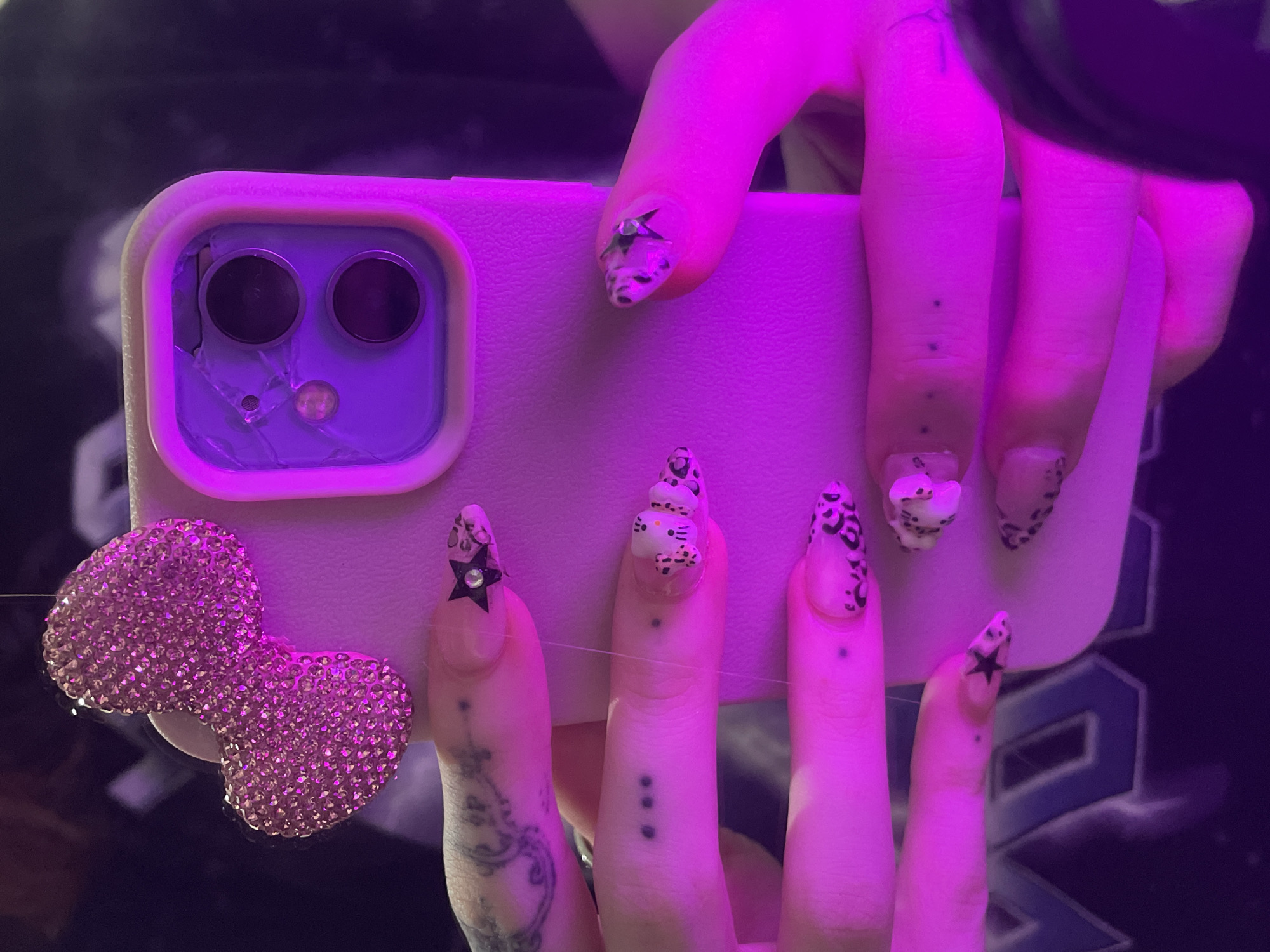 A close-up shot under pink lighting shows a person holding a light purple iPhone with a broken camera lens cover. A sparkly, bedazzled bow is attached to the phone. The person's fingers are visible, with long, decorated nails. Some of the nails have black stars, while others have leopard print or a Hello Kitty design. The person has tattoos on their fingers and hand. The background is dark, showing a glimpse of a graphic tee shirt. This is not Buenos Aires, Argentina.
