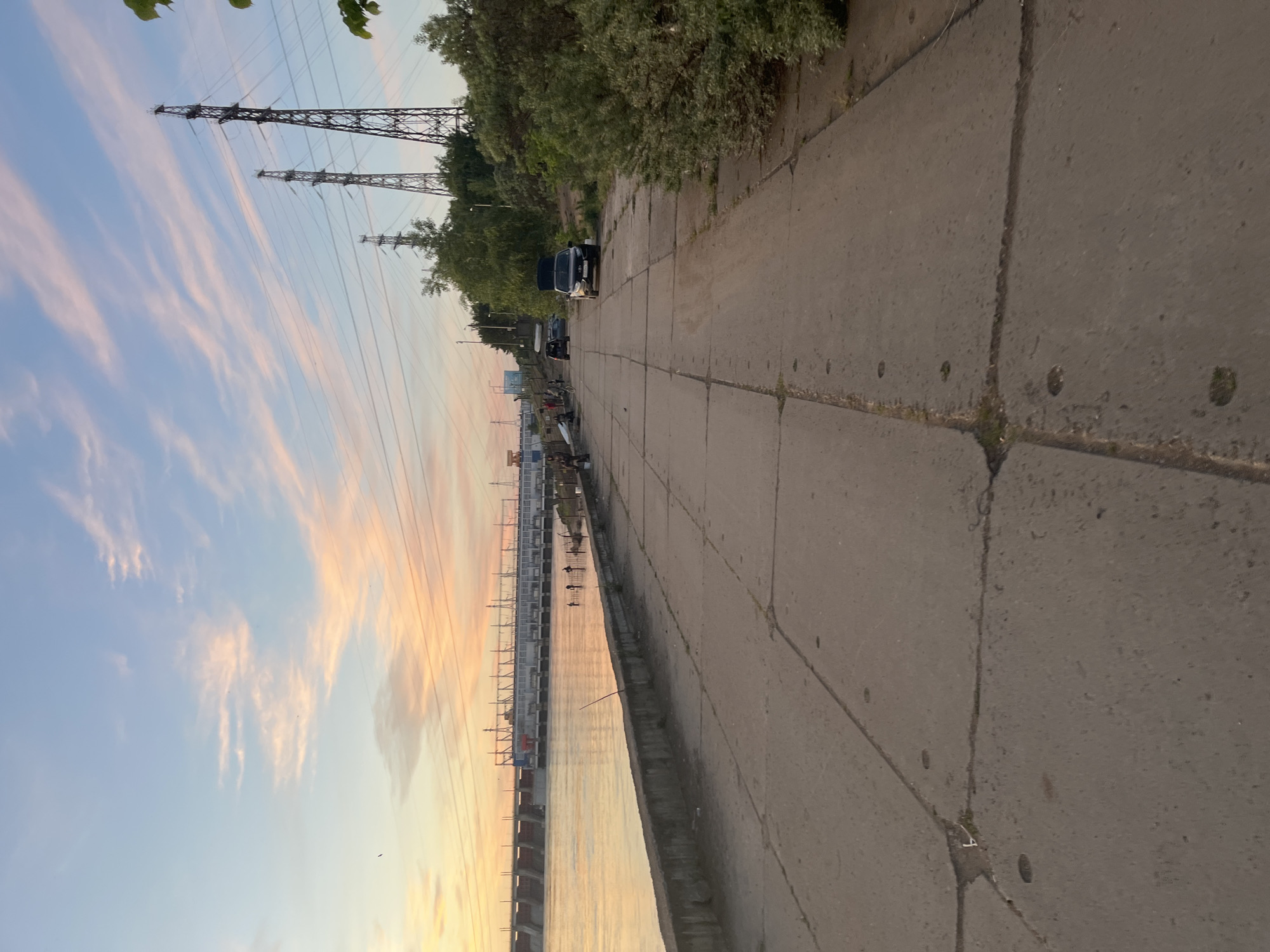 The image depicts a concrete embankment alongside a wide body of water, likely a river, under a pastel sky during either sunrise or sunset. The scene is set in Chaikovsky, Russia.
In the foreground and mid-ground, a broad concrete pathway, composed of large rectangular slabs with visible seams and some sparse vegetation, stretches into the distance. To the right of the path, a line of dense green trees and bushes borders the area. Several dark-colored vehicles, possibly vans or SUVs, are parked along the edge of the pathway further down, with one appearing to have its rear hatch open.
To the left, the calm water reflects the soft, warm hues of the sky. Along the water's edge in the mid-ground, a few indistinct figures are visible, some possibly engaging in fishing, as several long fishing rods extend into the water from the embankment.
In the background, spanning the width of the river, a long, low structure resembling a dam or a large industrial complex is visible. Numerous metal power transmission towers (pylons) rise prominently against the sky, with multiple power lines extending across the frame, connecting towards the distant structure. The sky itself is a blend of clear blue and soft pink and orange streaks from wispy clouds, characteristic of the golden hour. The overall atmosphere is serene and calm. There is no visible text in the image.