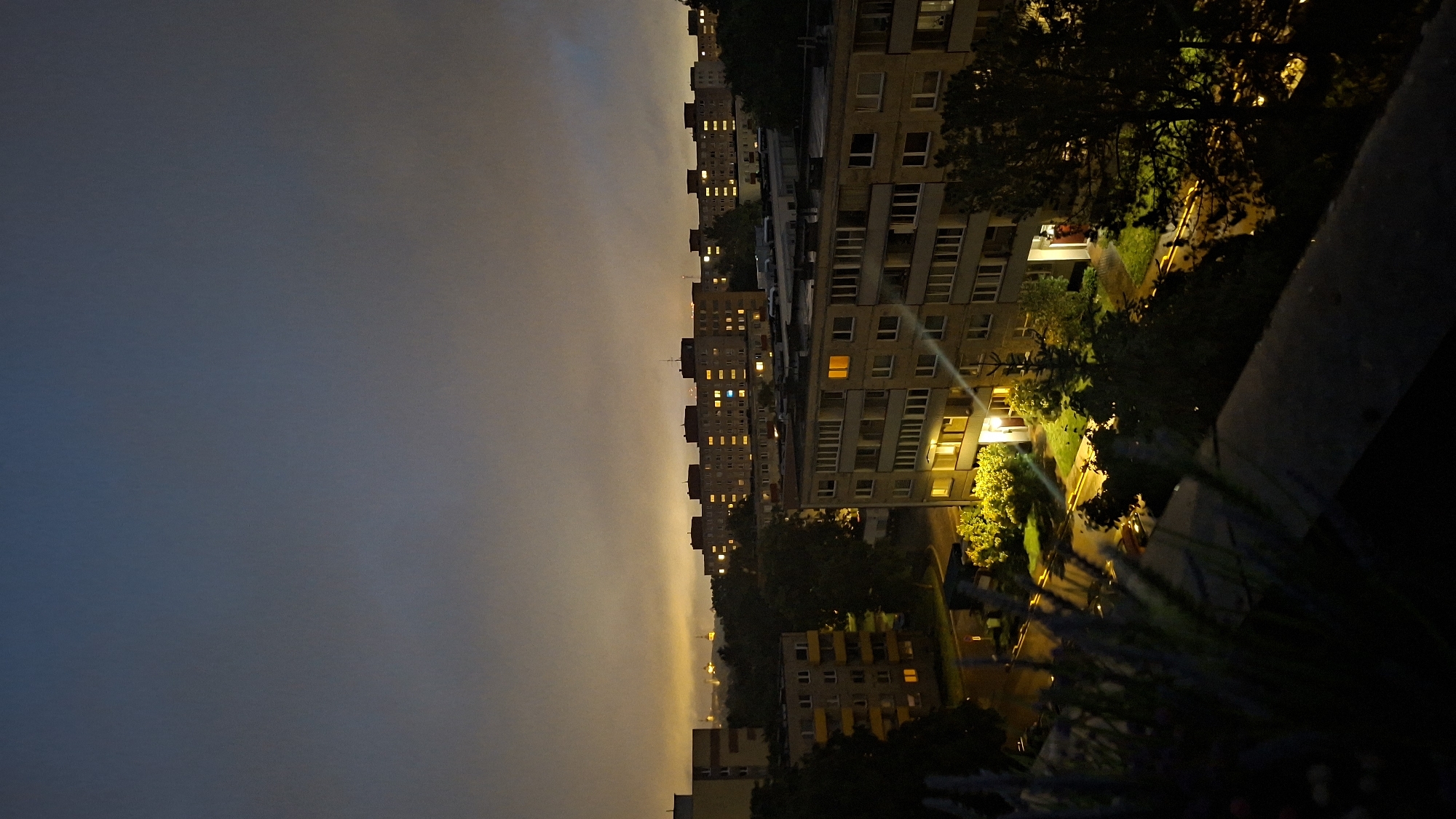 This is a high-angle, full-frame, vertical photograph taken at night in a residential area of Tallinn, Estonia. The scene depicts a view of apartment buildings and the sky. The sky is dark with a gradient from deep blue at the top to a hazy orange glow near the horizon. In the background, a distant city skyline with illuminated spires is faintly visible through the haze.
The foreground of the image is dominated by several apartment buildings. The closest building in the lower right and center is a multi-story concrete structure with numerous windows, many of which are illuminated with warm yellow light, indicating occupancy. Its facade is textured and shows signs of age. To the left of this building, there are trees and bushes, some of which are lit by artificial light sources, creating bright patches of green.
Further back, a series of long, block-style apartment buildings stretch across the middle ground. These buildings have a uniform design with multiple floors and a long, continuous row of windows. Many of these windows are also lit, creating a pattern of light against the dark structure. The buildings are set back from the immediate foreground, with a street or courtyard area visible between them and the foreground trees.
The overall impression is one of a typical urban residential landscape at night, with the artificial lights of the buildings contrasting with the darkness of the sky and surrounding vegetation. The presence of distant city landmarks suggests an urban setting within Tallinn. No people or specific actions are clearly discernible due to the low light conditions and distance.