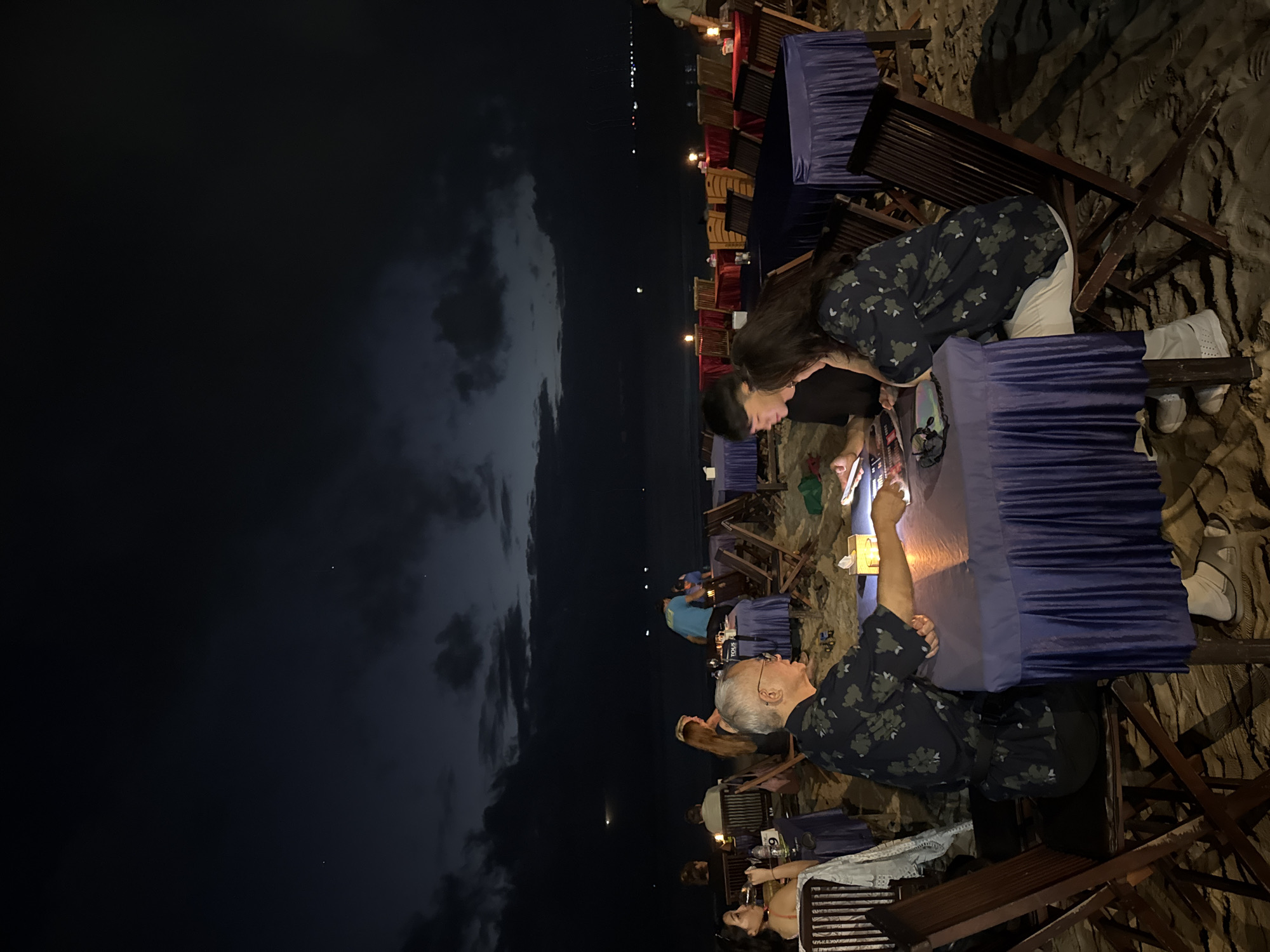 In Kuta Selatan, Indonesia, an outdoor beachside dining scene unfolds at dusk. Several tables covered with dark blue tablecloths are set up on the sandy beach, each with chairs around them. A group of people are seated at one of the tables in the foreground, engaged in conversation and looking at their phones. A man with glasses and a floral shirt is seated to the left, facing right, with his arm extended towards the table. A younger couple is seated opposite him, both looking down at a phone or tablet on the table. In the background, more tables with red and blue tablecloths are visible, some with lit candles, suggesting a restaurant or event. The sky above is dark and cloudy, with a faint horizon line of lights in the distance, possibly from other buildings or boats. The overall atmosphere appears relaxed and social, with a hint of romantic ambiance due to the low lighting and beach setting.