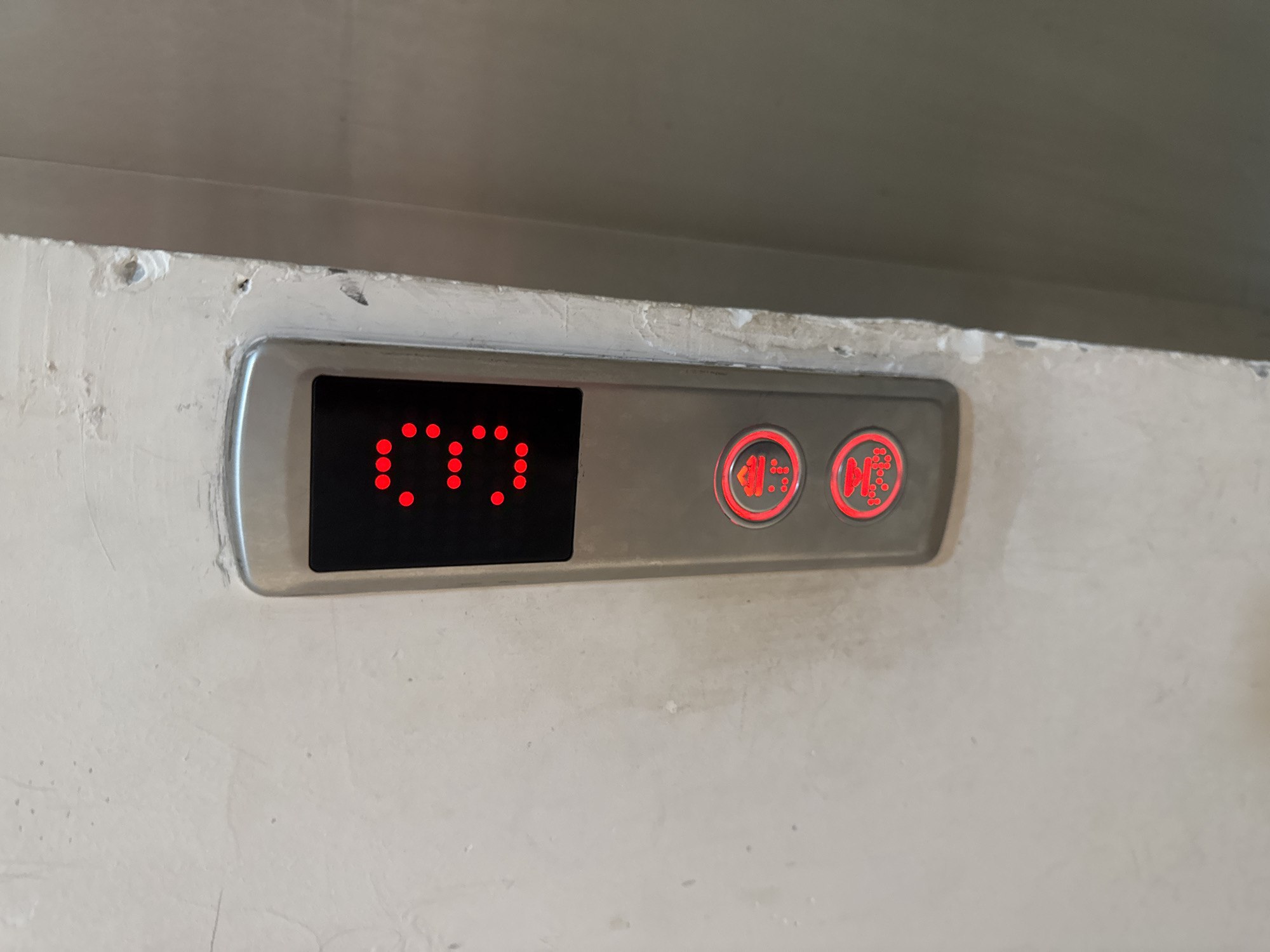 An elevator panel with a digital display showing the number "3" in red LED dots is mounted on a textured, off-white wall. Below the digital display are two red circular buttons, each with an icon and illuminated dots. The top button displays a simplified image of a person with an arrow pointing upwards, possibly indicating "up." The bottom button shows a similar person icon with an arrow pointing downwards, possibly indicating "down." The panel is made of brushed metal. The surrounding wall appears aged and slightly damaged, with visible imperfections. To the right of the panel is a darker, smooth surface, likely the interior of an elevator.