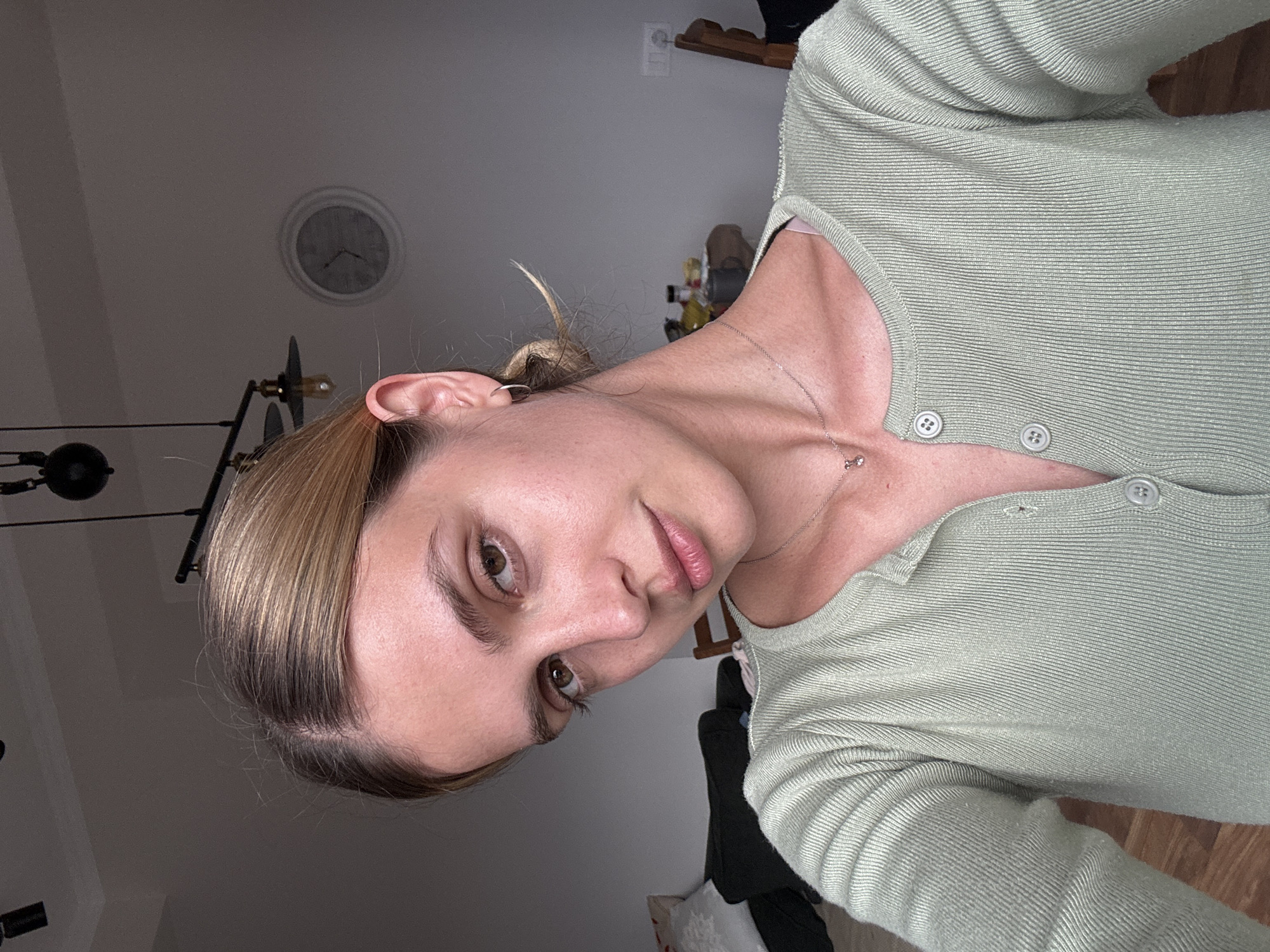 The image displays a close-up, angled view of a person, likely female, taking a selfie indoors. The person has light brown or blonde hair, pulled back, and striking brown eyes. Their complexion is fair, and they have a neutral expression with a slight pout on their lips.
They are wearing a light sage green, ribbed, long-sleeved top or cardigan featuring several small, light-colored buttons down the front. A delicate silver chain necklace adorned with a small, double-loop or heart-shaped charm is visible around their neck.
The background consists of light gray or off-white walls. In the upper left corner, a circular wall clock with dark hands is faintly visible, though the precise time cannot be determined, it appears to be around 12:45 or 1:00. A modern black metal lighting fixture with exposed bulbs is positioned on the left side of the frame. Towards the upper right, a wooden shelf holds indistinct items. A portion of a wooden floor is visible in the lower right, alongside some dark fabric, possibly furniture or a blanket.
The image is rotated approximately 90 degrees clockwise, suggesting it was taken with the camera held sideways or has been rotated post-capture. The overall lighting indicates an indoor setting, possibly daytime or a well-lit interior. There is no visible text.
