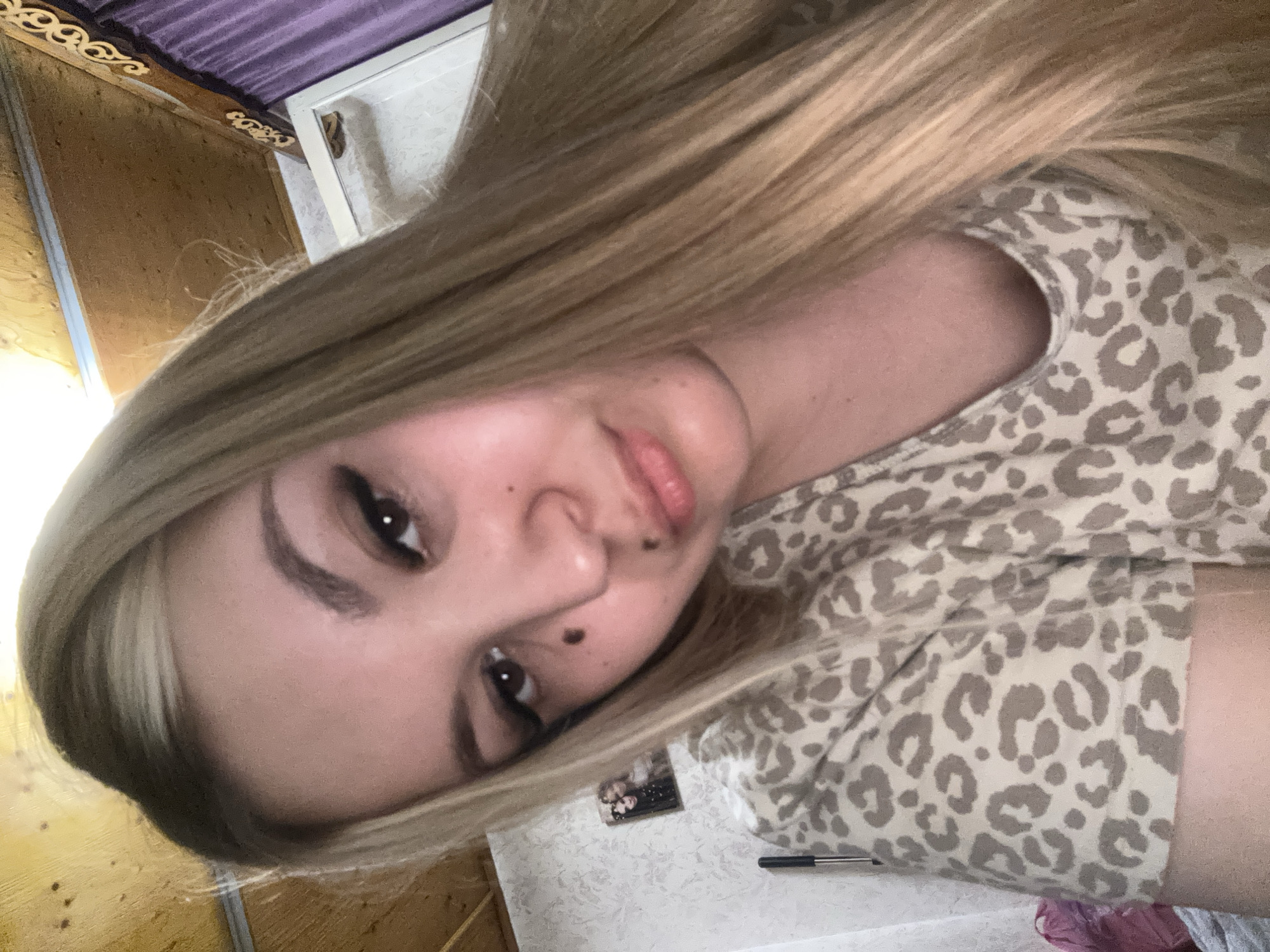 A selfie taken indoors shows a young Caucasian woman with long blonde hair and a leopard-print shirt. She has makeup on her eyes and lips. She is looking directly at the camera. The background includes a wall with a picture frame, a curtain, and a wooden ceiling. The scene appears to be a bedroom or living area. There is no specific information to determine the exact location within Большой Букор, Russia.