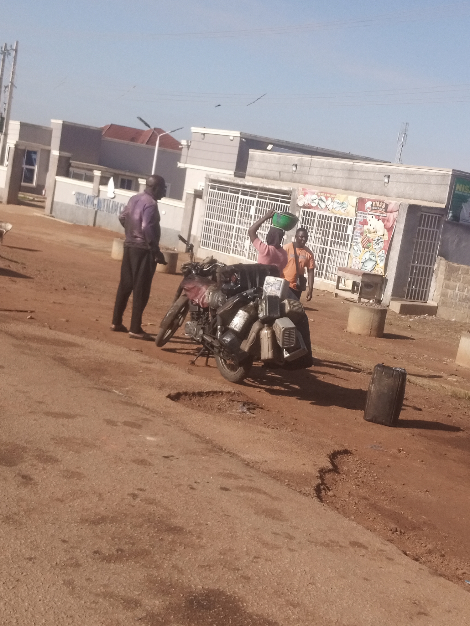 In an outdoor environment under a clear blue sky, a scene unfolds on a dirt and asphalt road. The foreground features a motorcycle laden with containers, parked on the side of the road. To the left, a man stands beside the motorcycle. Further back, two individuals are visible near a building. One person, a woman, carries a green bucket on her head, while the other, a man, stands beside her. The building in the background appears to be a commercial establishment with a white facade and barred windows. A poster is displayed on the building's exterior. The ground is a mixture of reddish-brown dirt and sections of asphalt, with some patches of uneven terrain. The overall atmosphere suggests a public space within a city, likely a marketplace or commercial area.