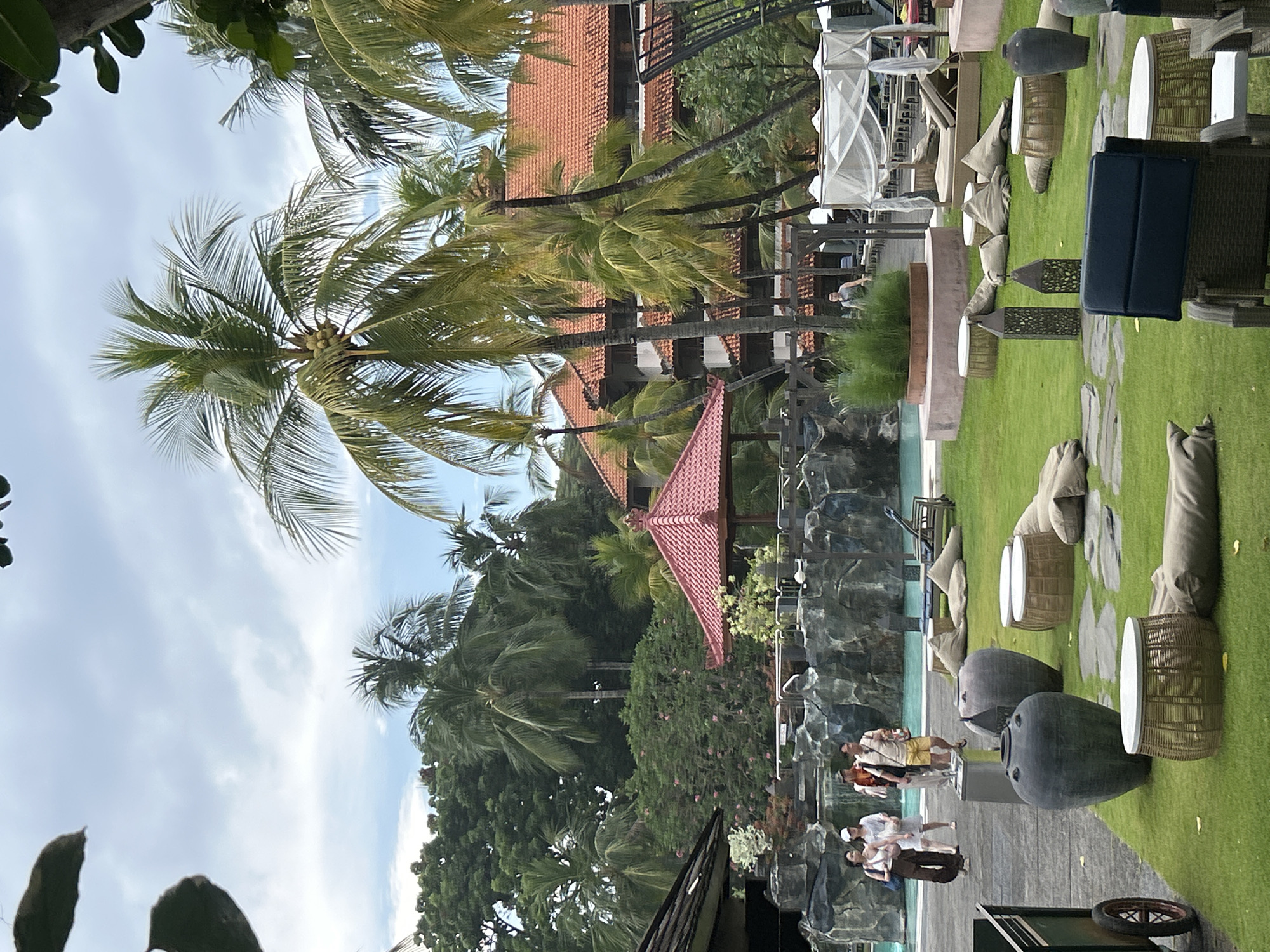 The image depicts an outdoor resort area with lush green grass, palm trees, and a swimming pool. Several people are walking near the pool, which has a rocky waterfall feature. Various seating arrangements are scattered across the lawn, including beanbag chairs, low tables with woven bases, and large decorative vases. In the background, a resort building with an orange tiled roof and several smaller structures are visible. The sky is cloudy. This scene is consistent with resort settings found in Kuta, Indonesia, known for its beaches and tourist accommodations.