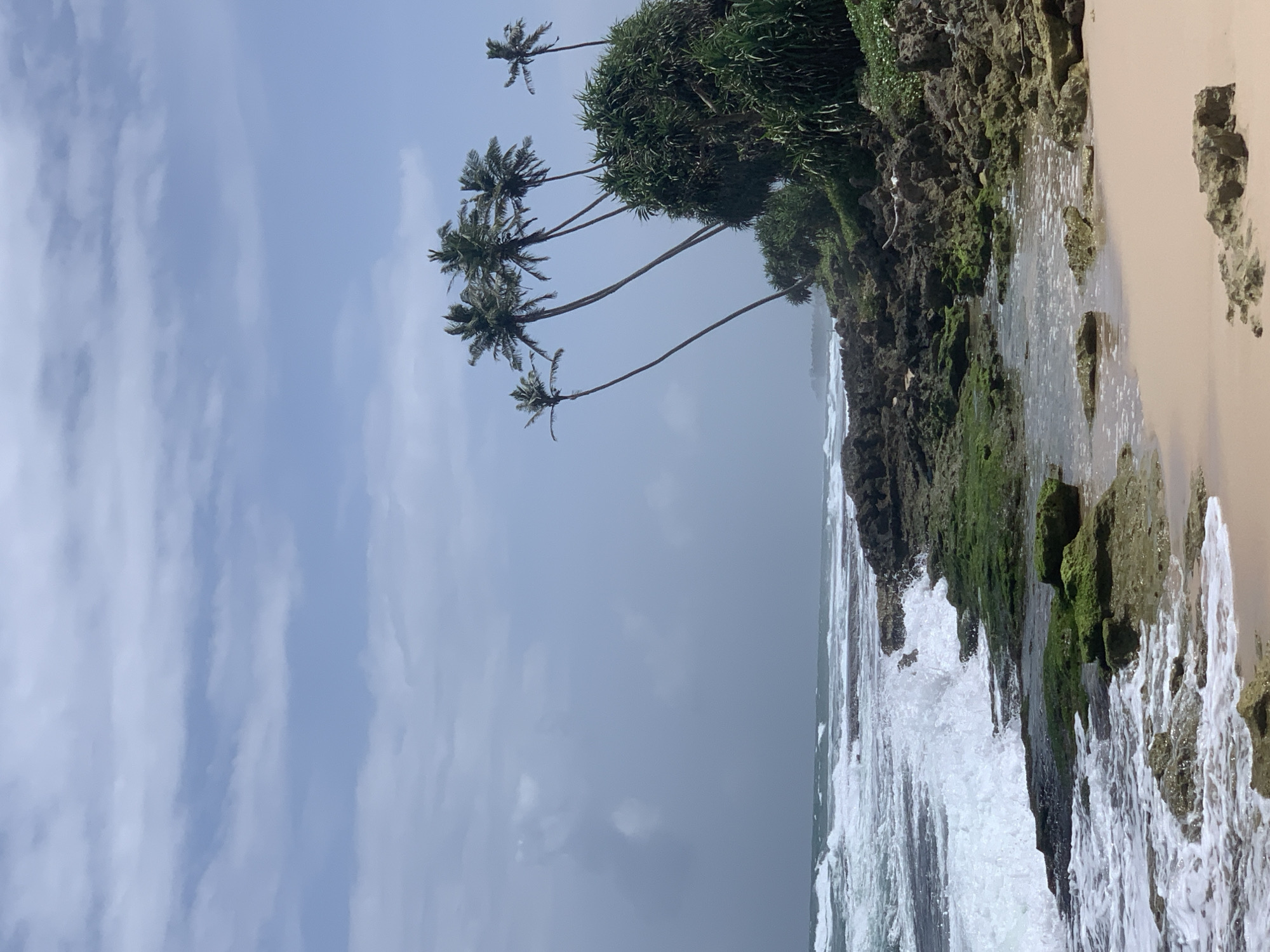 This image captures a picturesque coastal scene in Habaraduwa, Sri Lanka. The focal point is a rocky shoreline with waves crashing against it, creating white foam. Lush green vegetation, including palm trees, grows along the top of the rocks, adding to the tropical feel. The ocean stretches out to the horizon, meeting a sky filled with soft, white clouds against a pale blue backdrop. The beach to the right is a light, sandy color.
The image appears to have been taken during the daytime, with the lighting suggesting it could be morning or late afternoon, as the sky is not intensely bright. The weather seems mild and possibly overcast or nearing a storm, given the muted sky colors and the dramatic waves. The presence of palm trees and the rocky coastline are strong indicators of a tropical, island location.
No people or specific objects are prominently featured, emphasizing the natural beauty of the landscape. The overall mood is serene and wild, showcasing the power of nature where the land meets the sea. There is no visible text in the image.