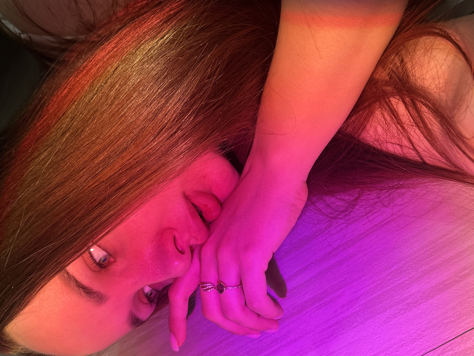A woman with long, reddish-brown hair is lying down, her face close to the camera. Her right hand, adorned with two rings, is near her mouth. The background is a surface with a wood-like texture. The lighting casts a red and purple hue across the scene. There is no visible information to confirm the image was taken in Chaikovsky, Russia.